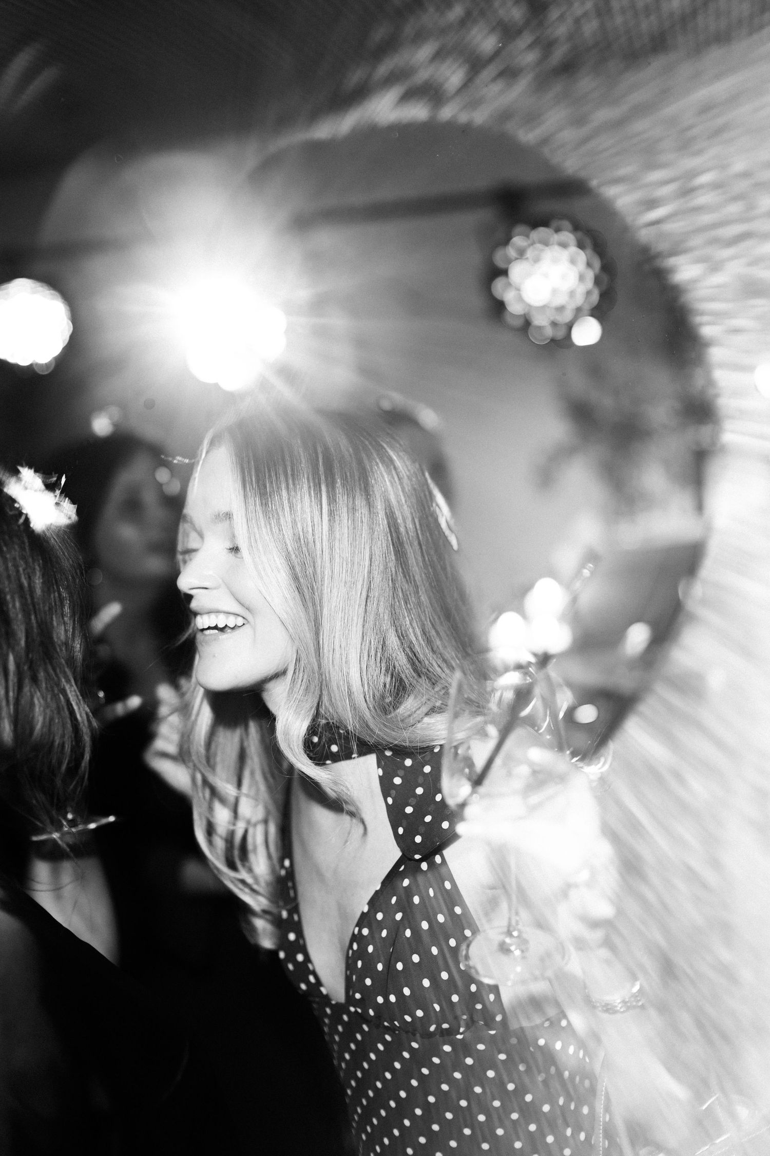 Black and white photo of a smiling woman in polka dot dress dancing at a party with bright lights.
