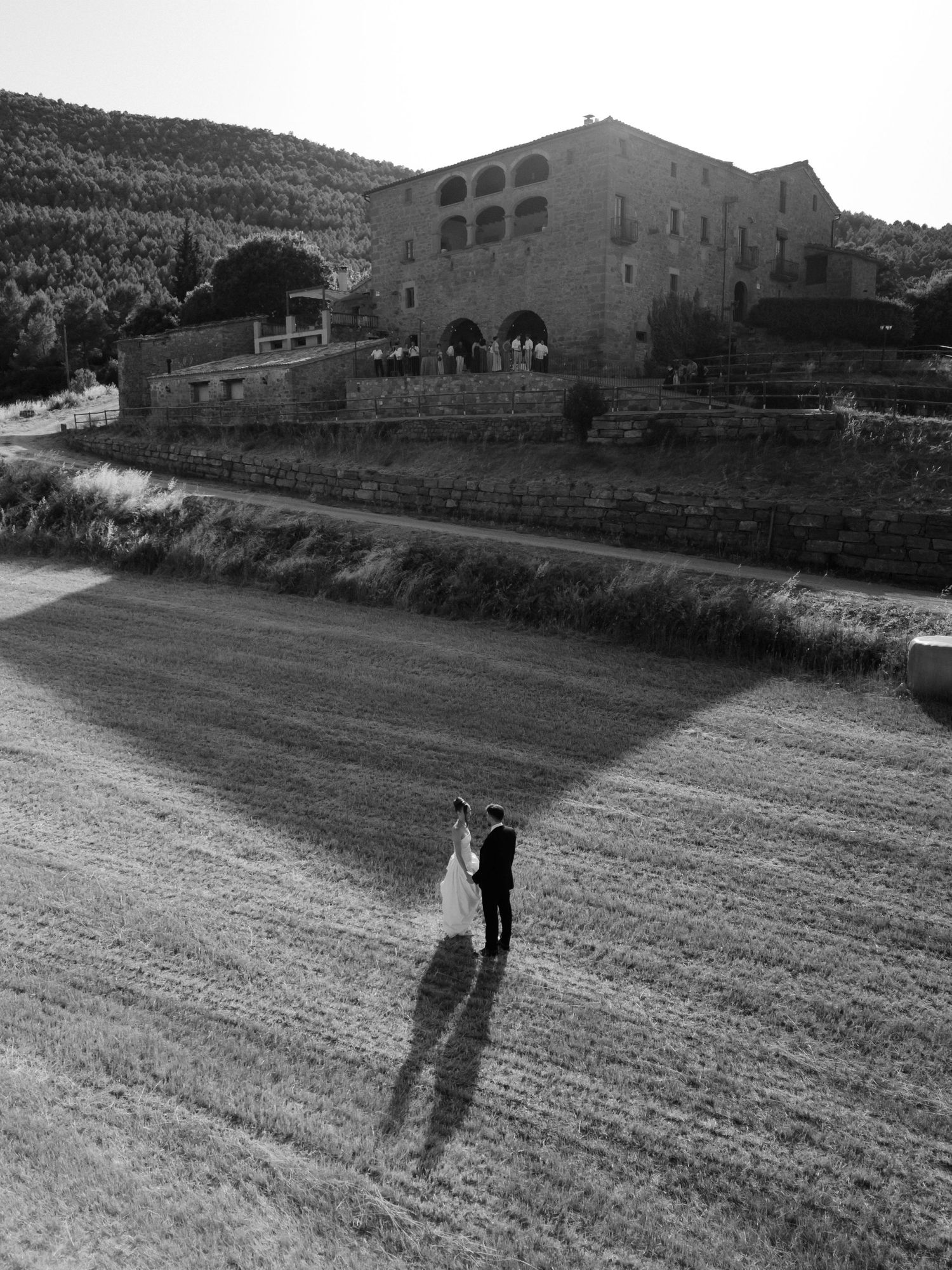 Black and white aerial photo of a bride and groom embracing in a field beside a historic stone villa in the countryside.