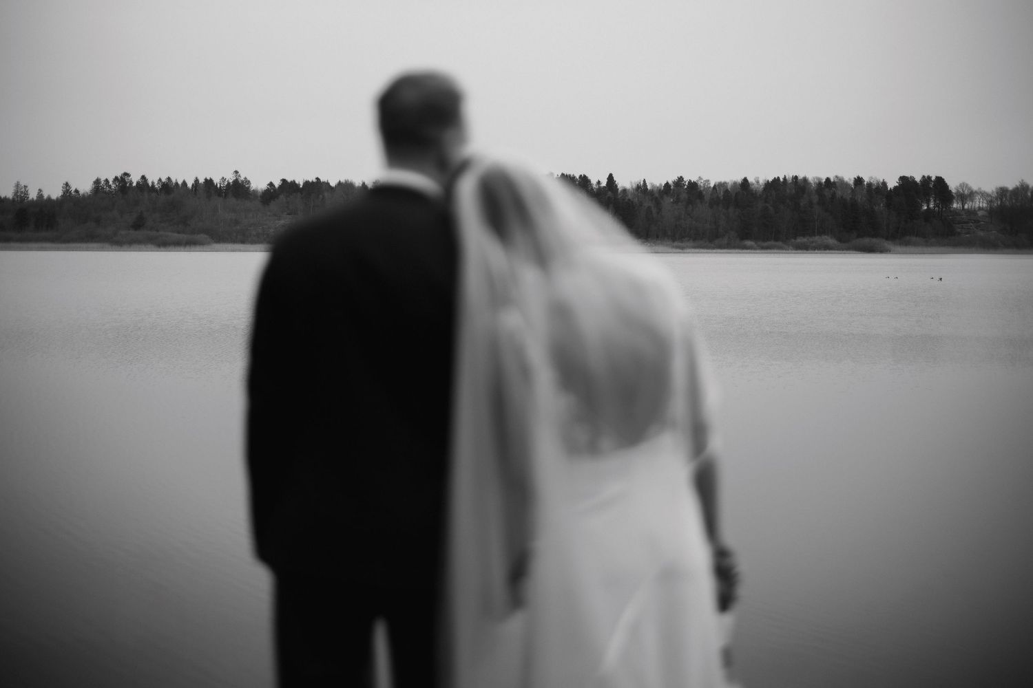 Black and white photo of bride and groom standing together gazing at a calm lake.