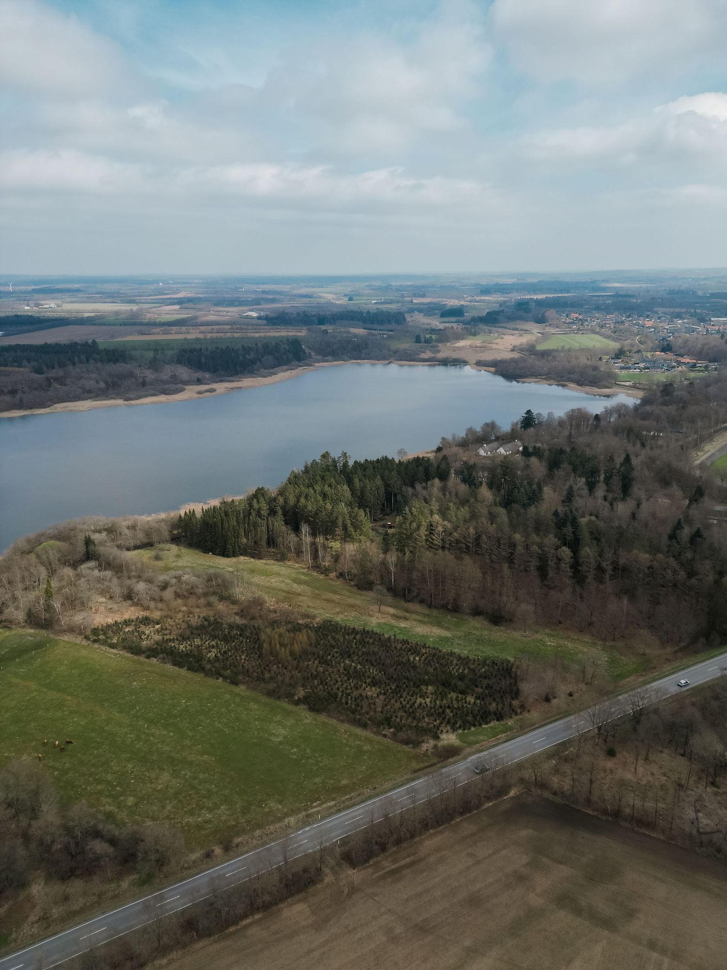 Aerial view of a large lake surrounded by forests and farmland under a partly cloudy sky.