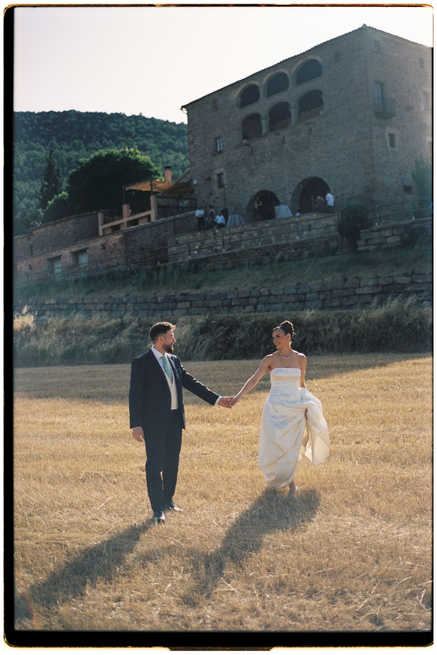 A bride and groom walk hand-in-hand through a golden field near a rustic stone villa at sunset.