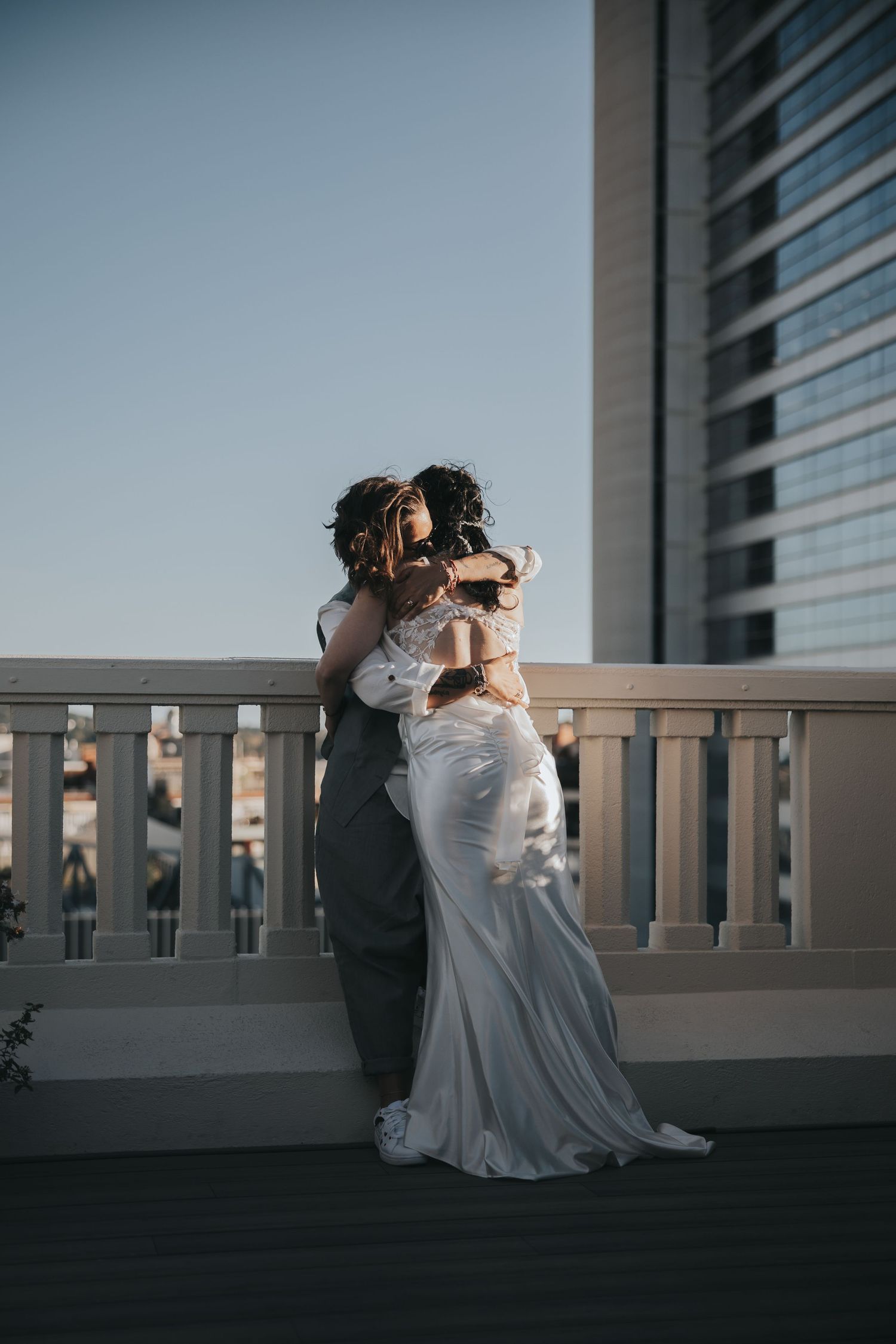Bride and groom embracing on a rooftop balcony at sunset with a city skyscraper in the background.