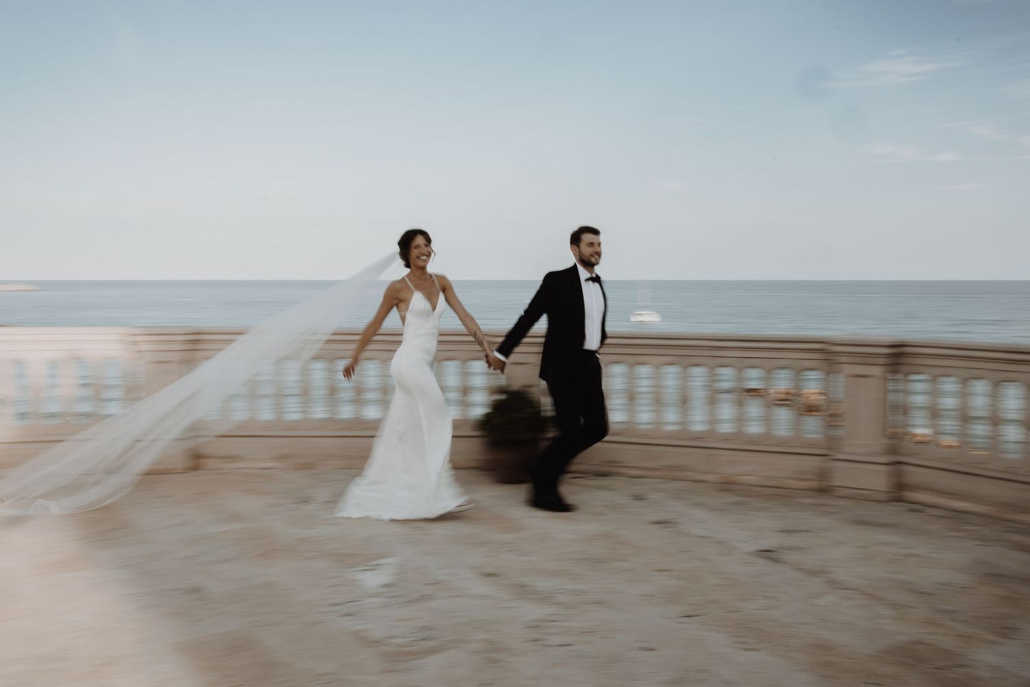 Bride and groom holding hands walking on a seaside terrace with ocean views on their wedding day.