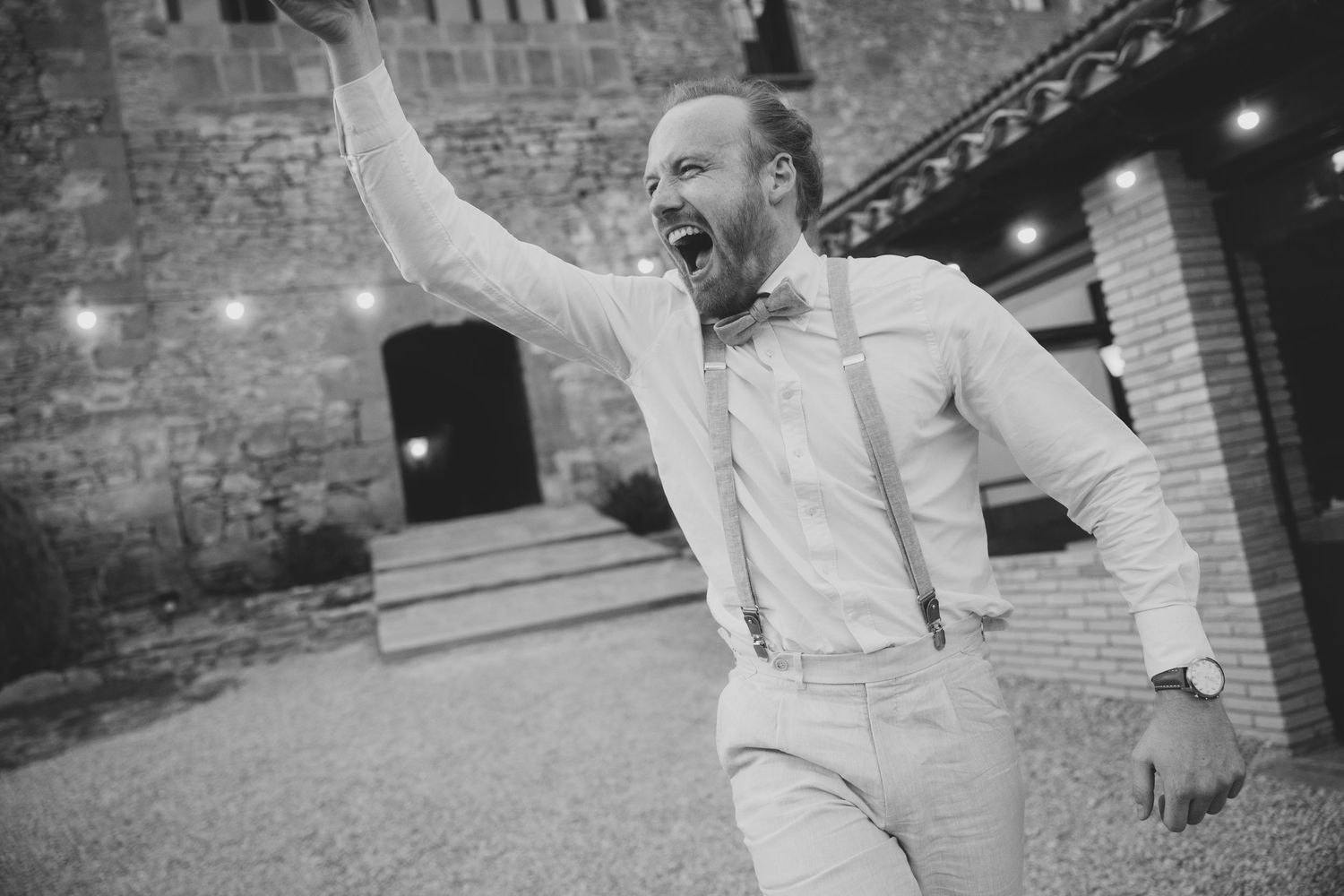 Black and white photo of an excited groomsman in suspenders celebrating outdoors at a wedding venue.