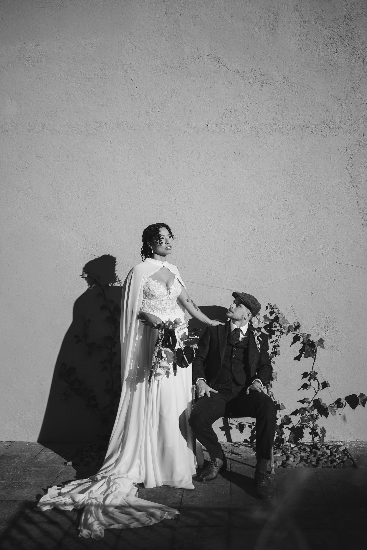 Black and white photo of bride in cape gown and groom in suit posing against a textured white wall.