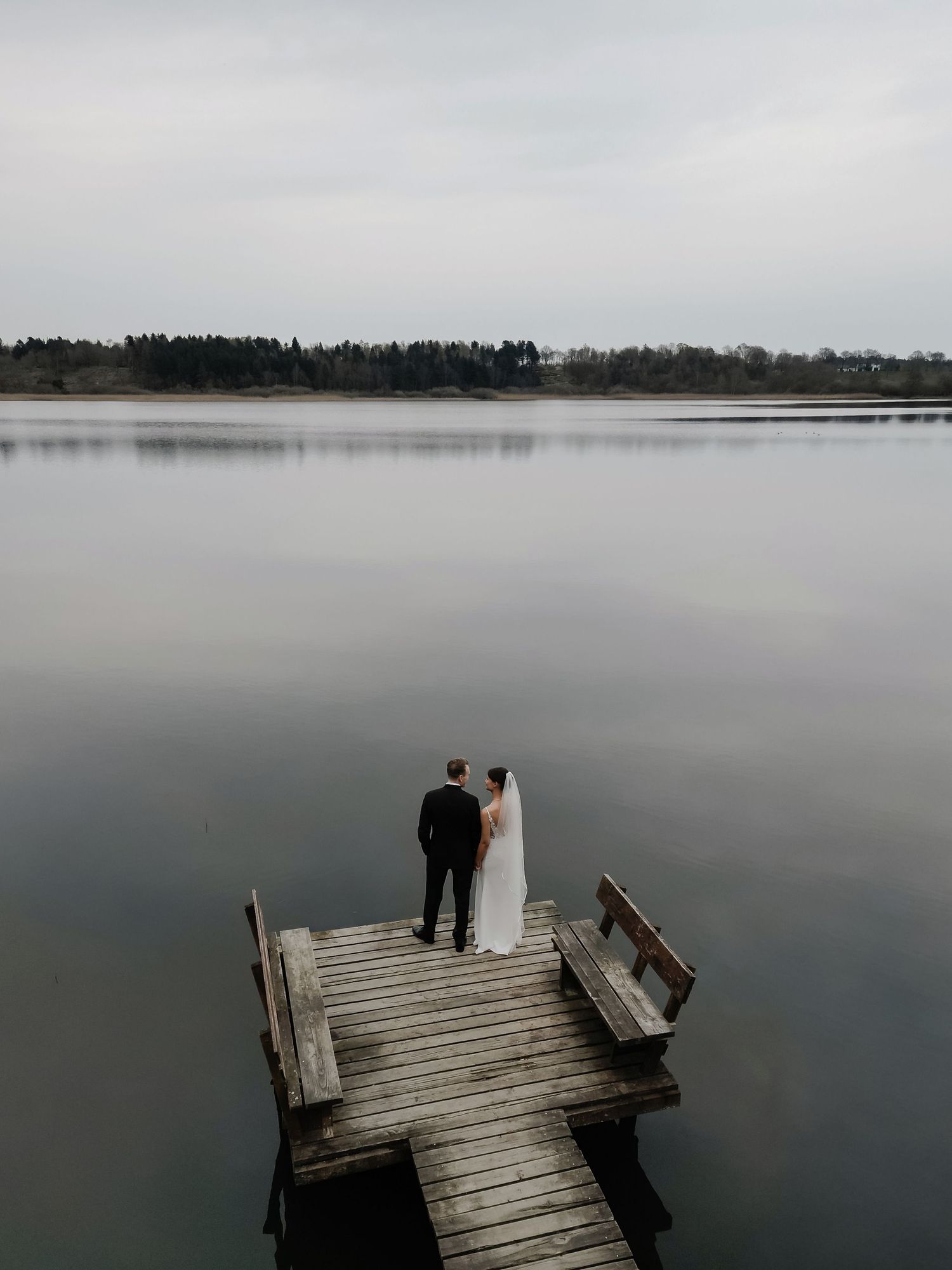 Bride and groom standing on wooden lakeside dock under overcast sky with calm water.