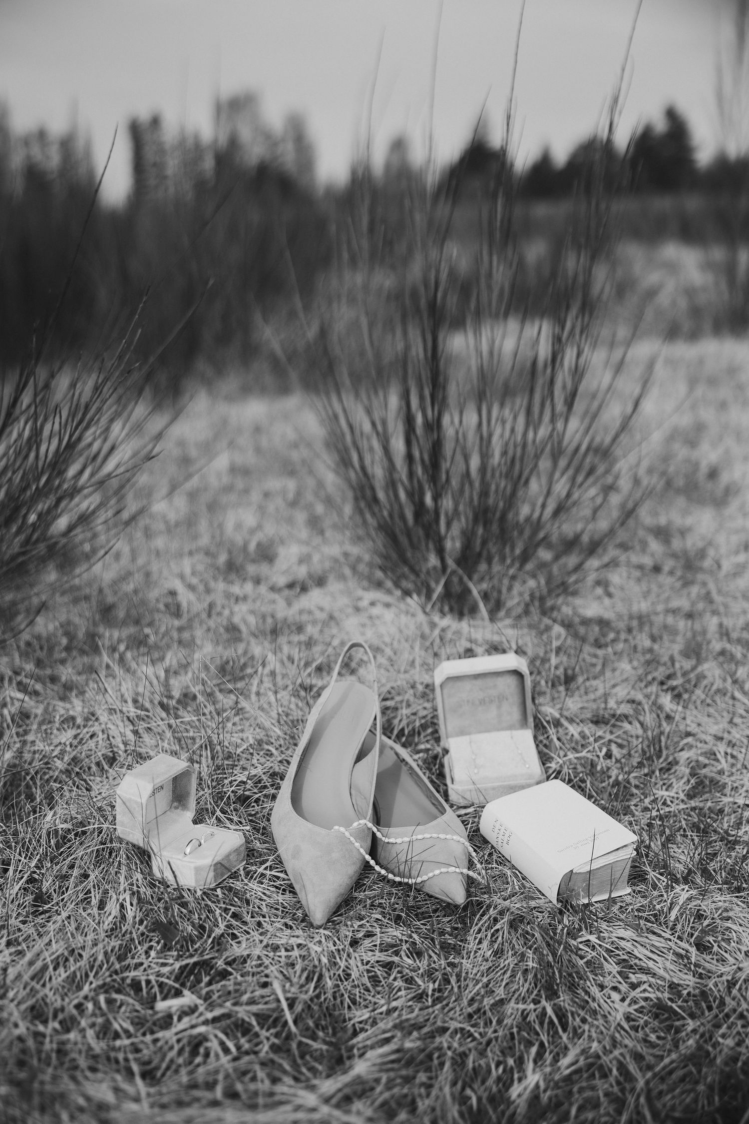 Black and white photo of bridal shoes and ring boxes arranged on grass in a rustic outdoor field setting.