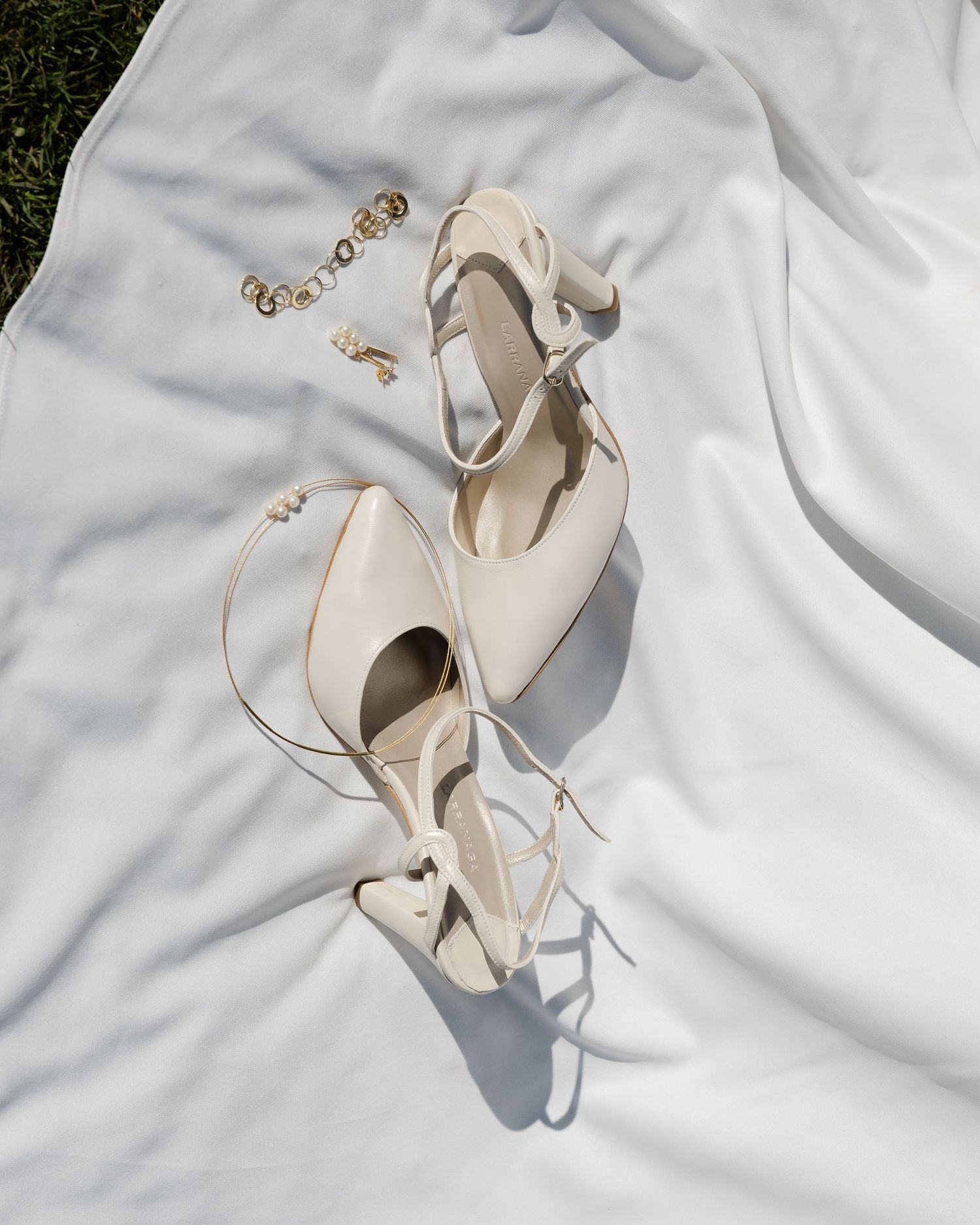 Elegant ivory bridal heels with ankle ties and gold jewelry on white fabric.
