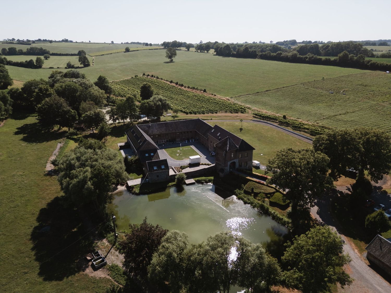 Aerial view of a historic moated manor house surrounded by lush green fields and trees on a sunny day.