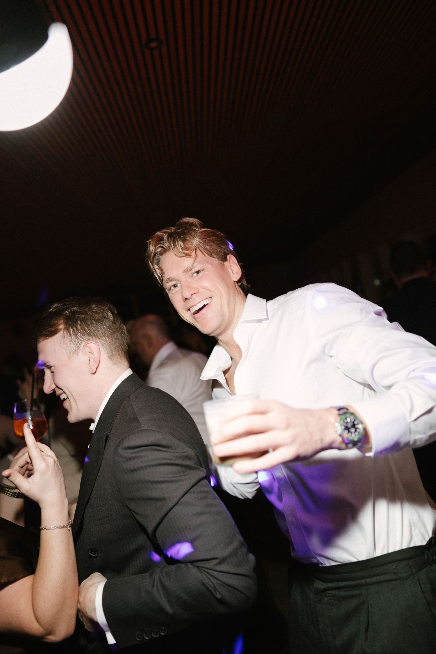 Two men laughing and dancing at a party, one holding a drink under purple lights.