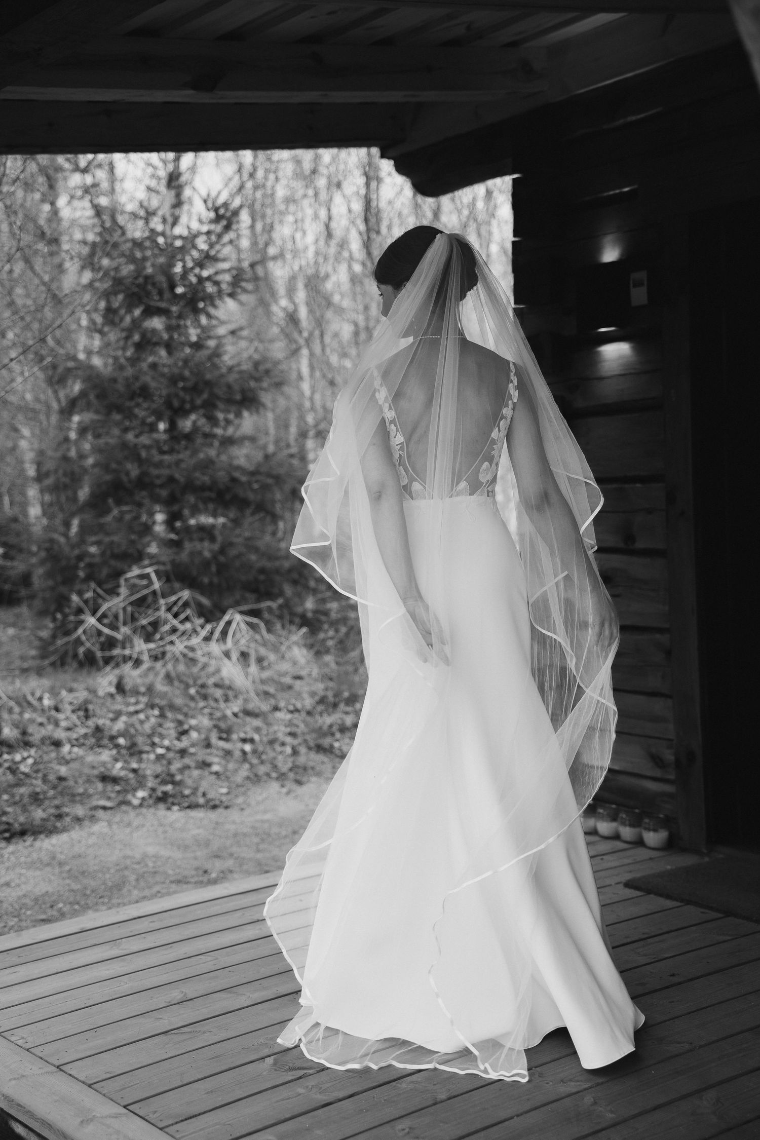 Bride in elegant white wedding gown and layered veil standing on wooden deck surrounded by forest.