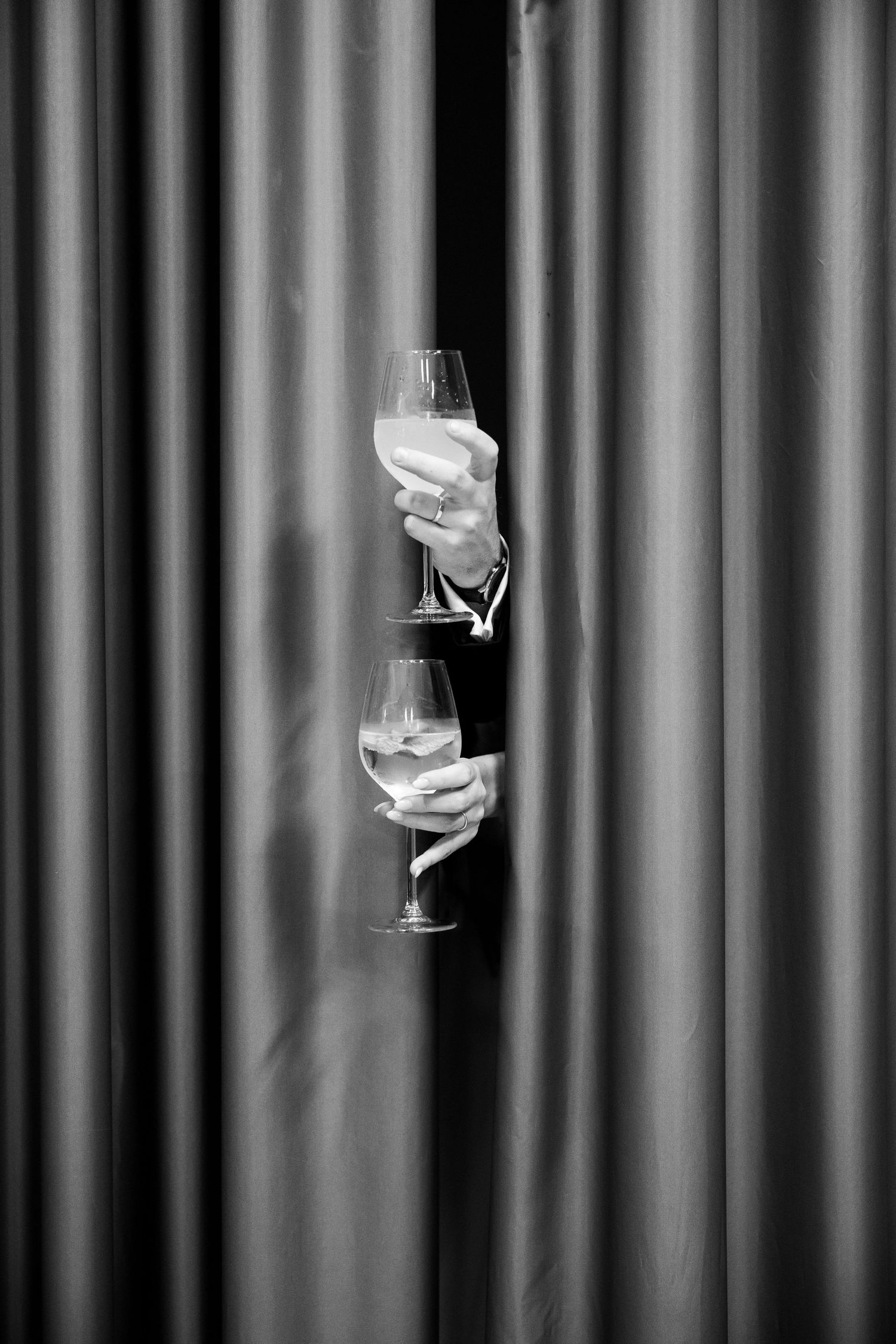 Two hands emerging from curtains holding wine glasses in elegant black and white photography.
