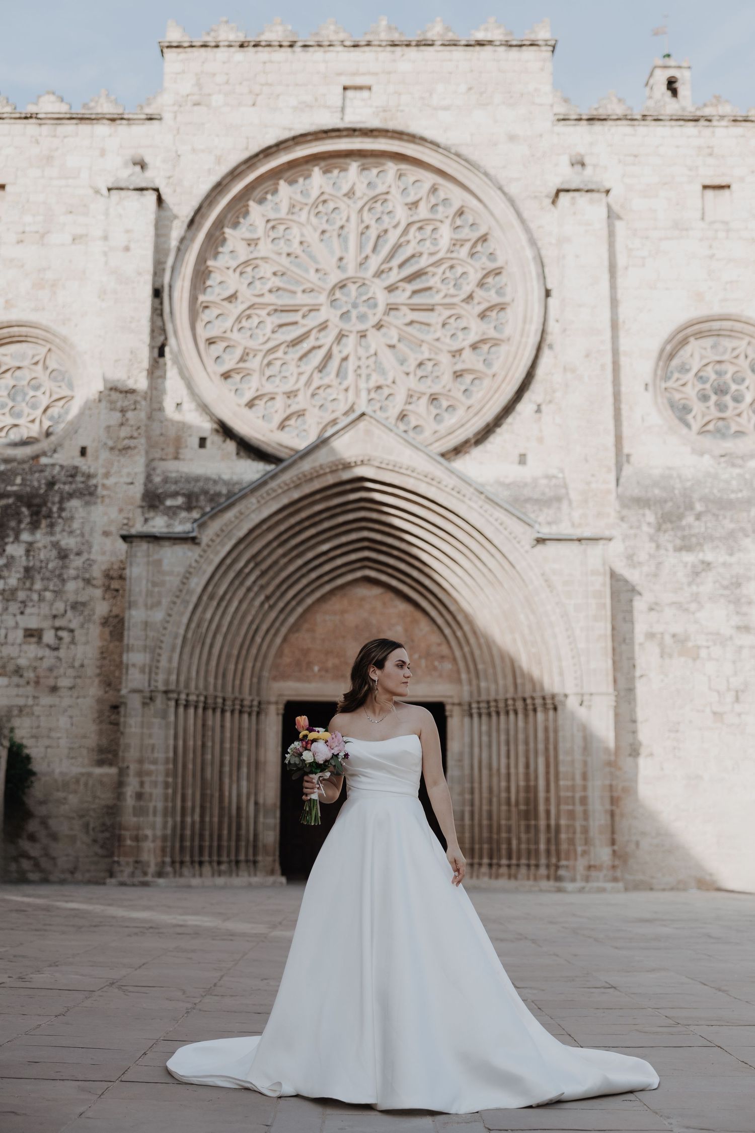 Bride in white off-shoulder gown standing before a Gothic cathedral with rose window.