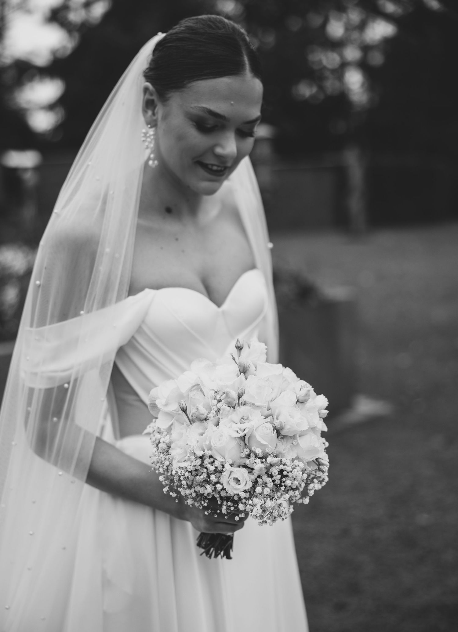 Elegant bride in strapless gown and pearl veil holding a bouquet of roses and baby's breath, black and white photo.