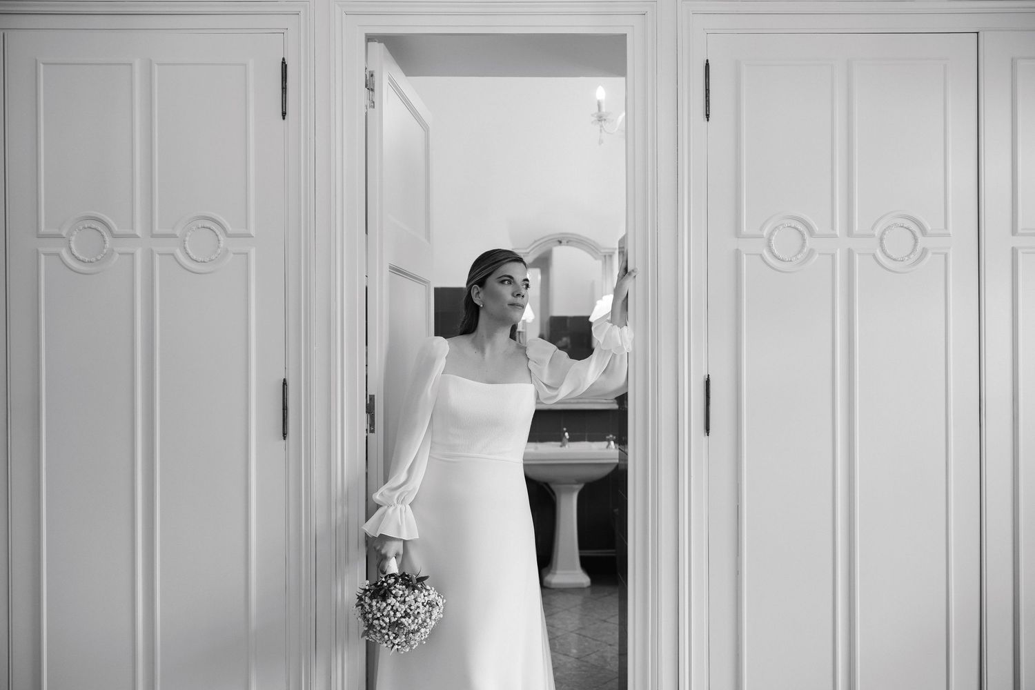 Black and white photo of bride in long-sleeve gown holding bouquet in ornate white doorway.