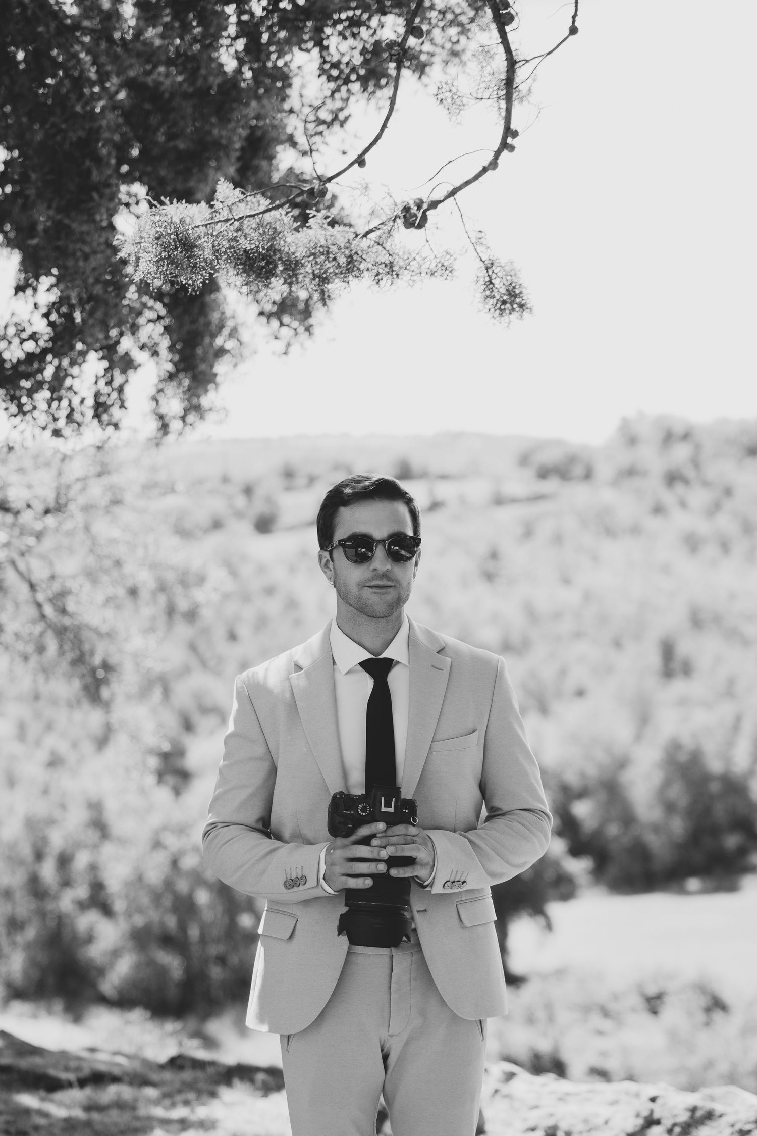 Elegant man in light suit and sunglasses holding a camera outdoors in a black and white portrait.
