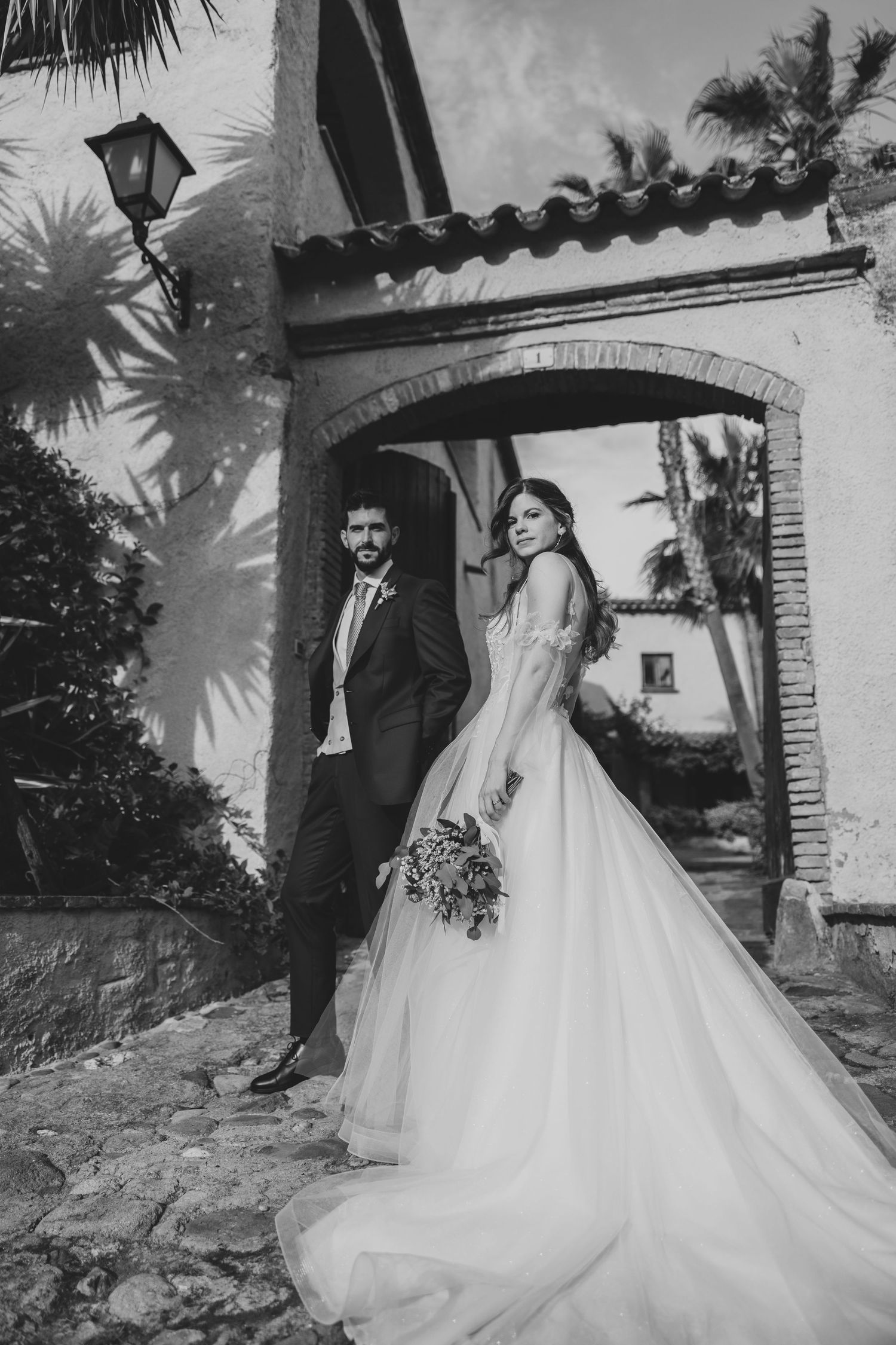Black and white photo of bride in ballgown and groom standing under a brick arch with palm trees.