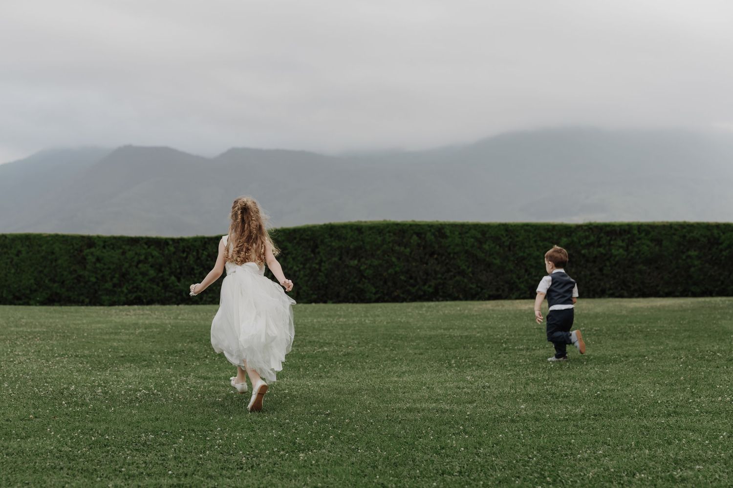 Bride in white dress runs across green lawn with young boy, mountains visible in misty background.
