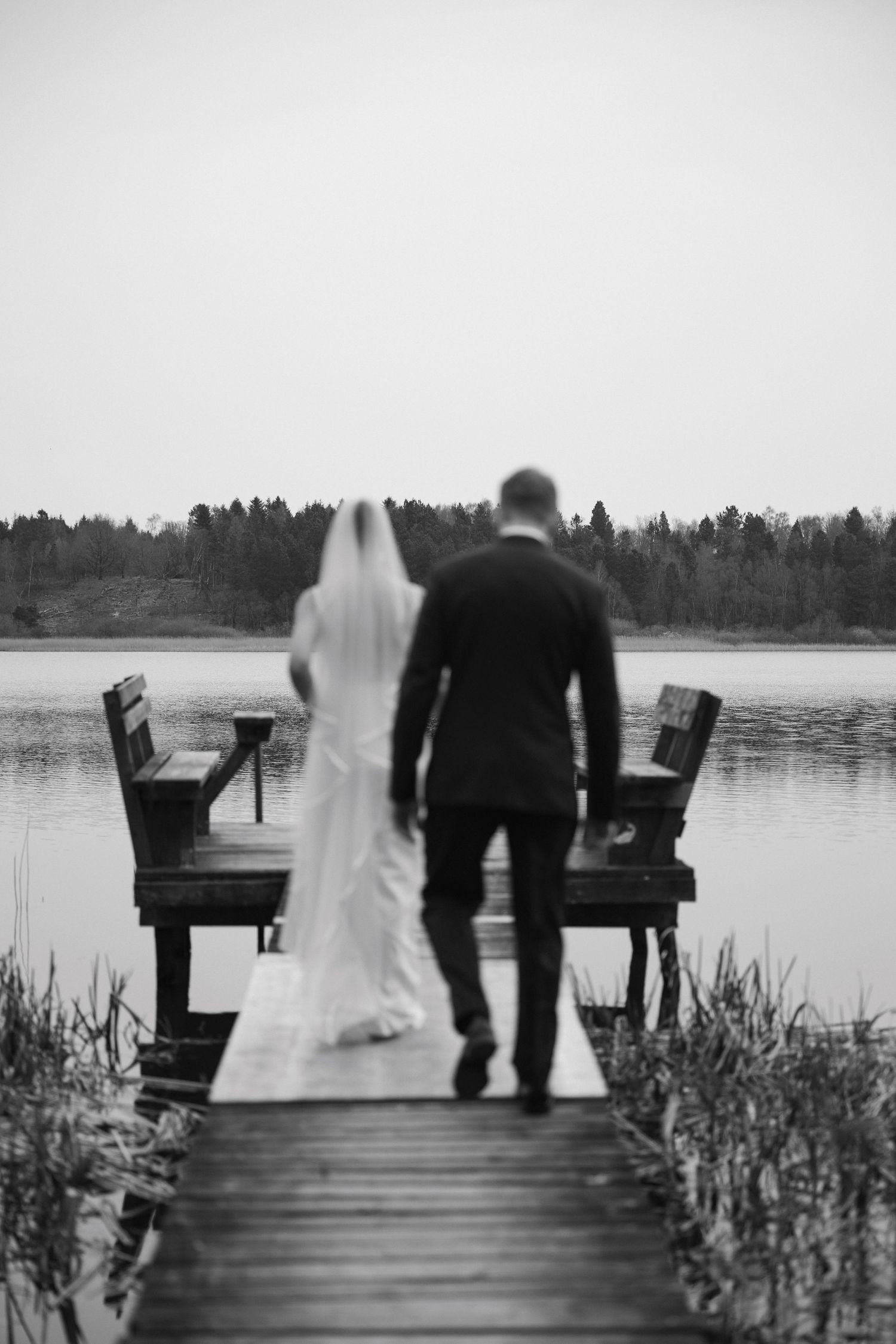 Black and white photo of a bride and groom walking hand in hand on a wooden dock overlooking a calm lake.