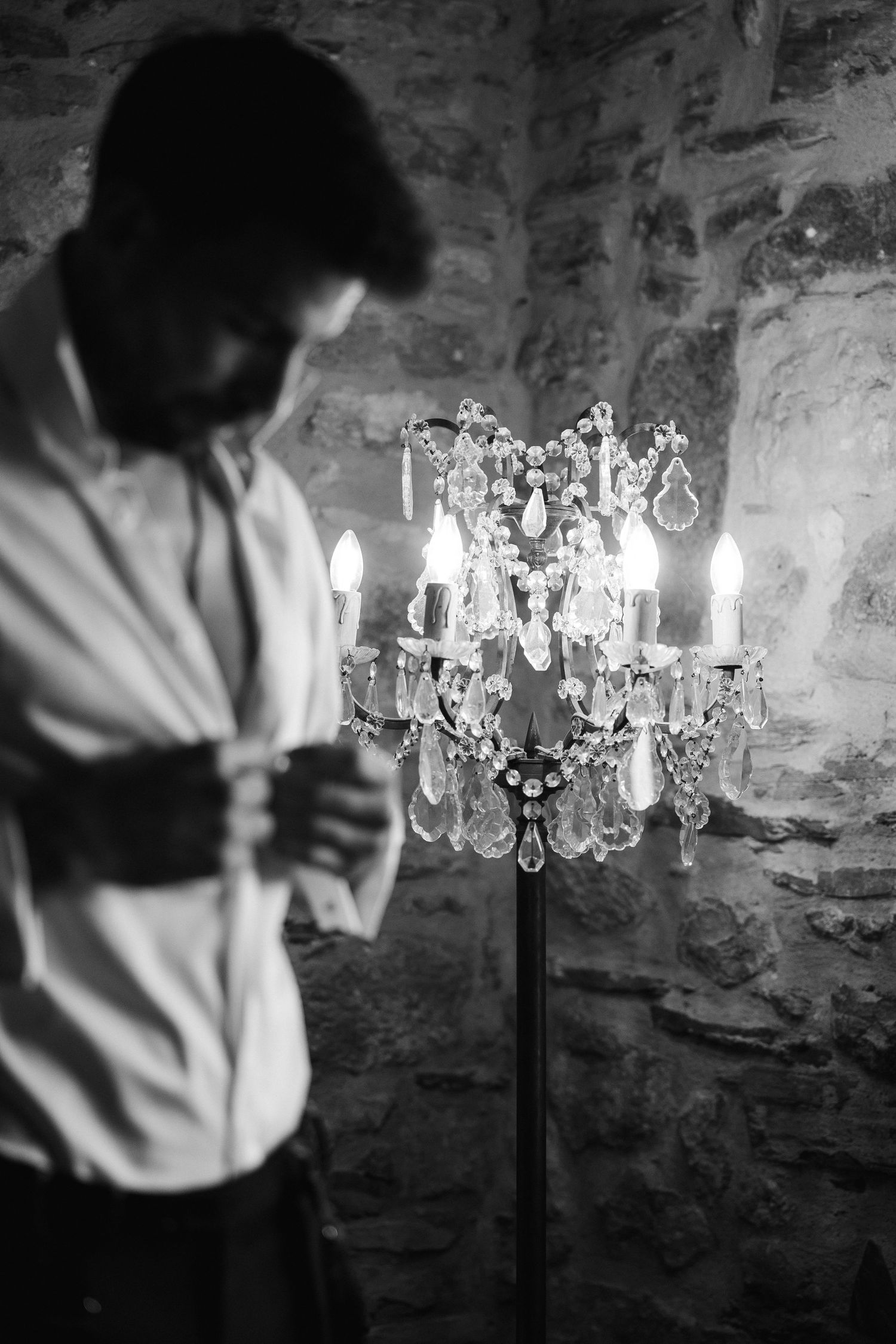 Black and white photo of bearded man in white shirt beside elegant crystal candelabra against stone wall.