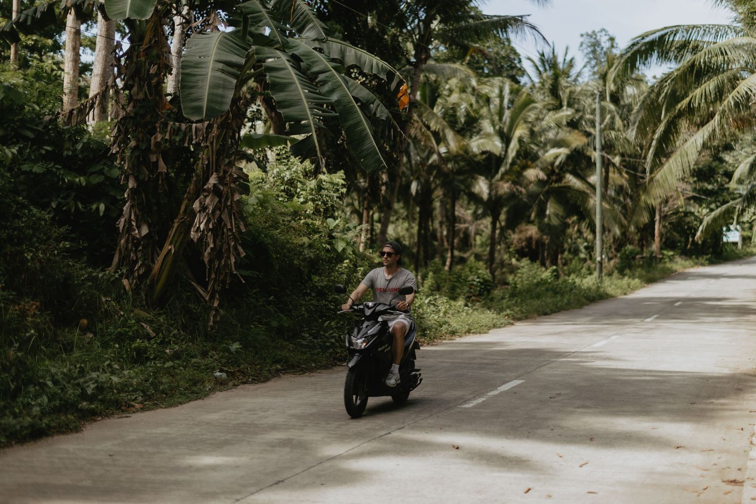 A man rides a motorcycle along a tropical road lined with lush palm and banana trees.