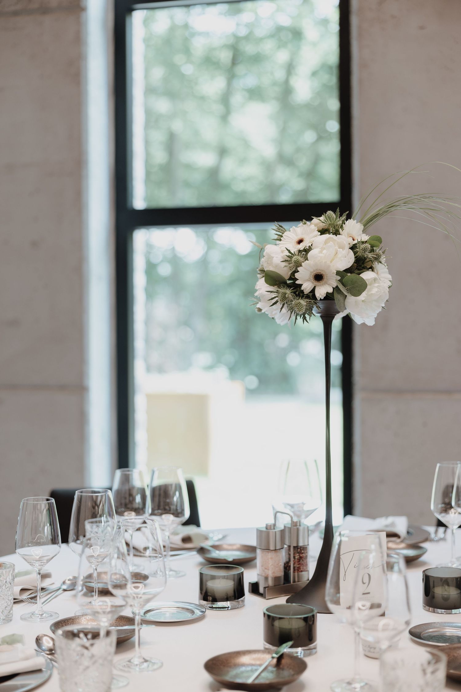 Elegant wedding reception table with tall white floral centerpiece, crystal glasses, and large window.