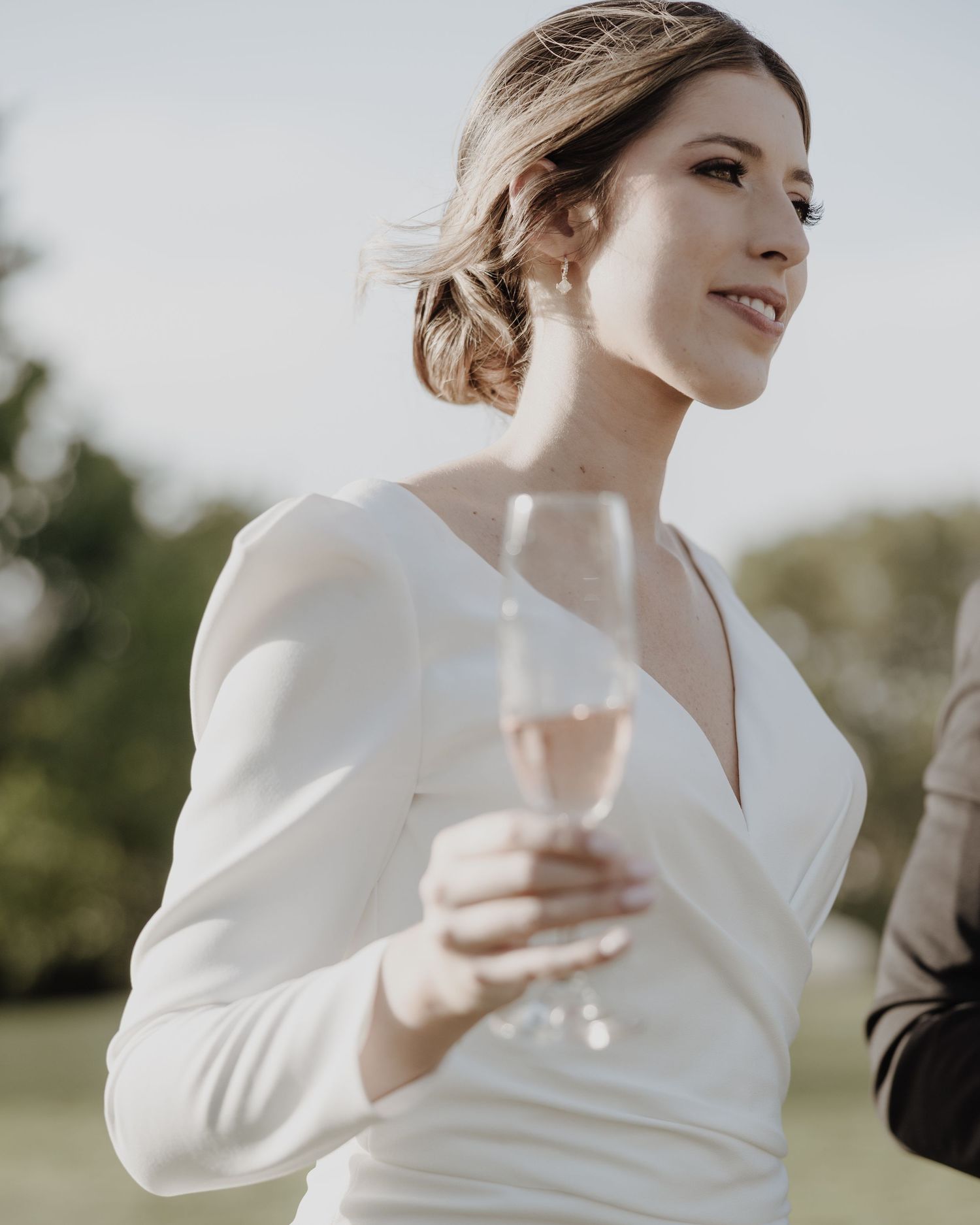 Beautiful bride in white long-sleeve wedding dress holding champagne glass outdoors at golden hour.