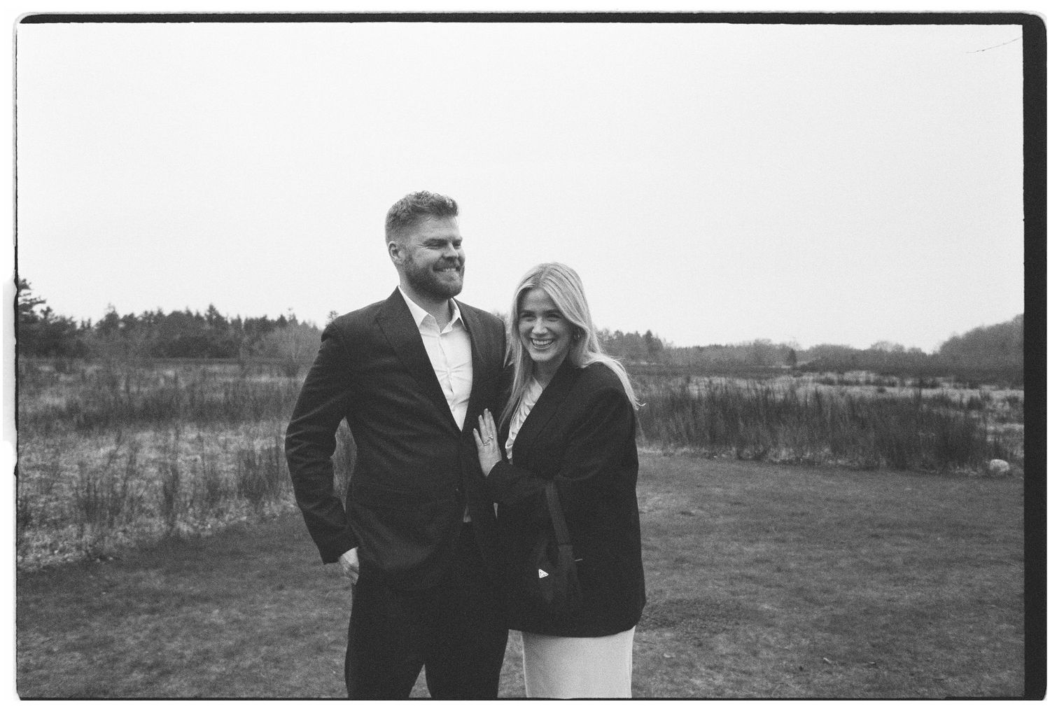 Smiling couple in dark outfits posing together in an open field in black and white film style.
