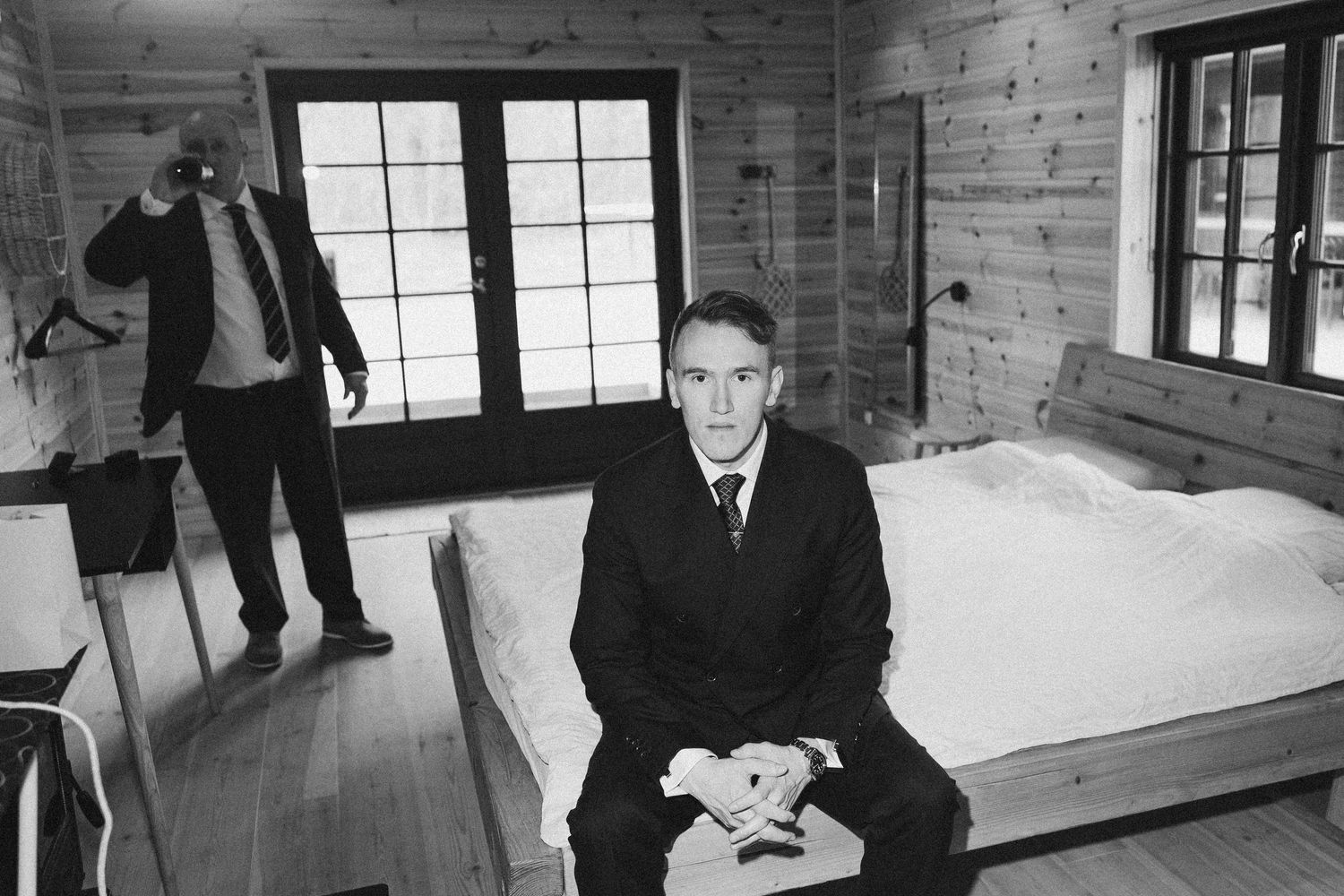 Black and white photo of two men in suits inside a rustic wooden cabin bedroom.