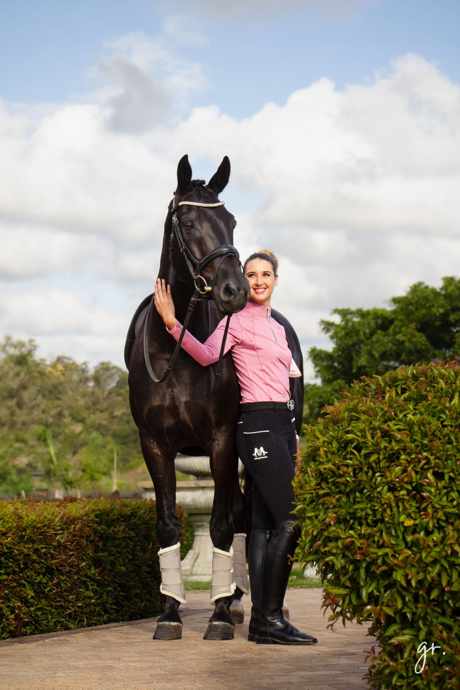 A black horse stands next to someone wearing a pink top and black pants in an outdoor setting with greenery.