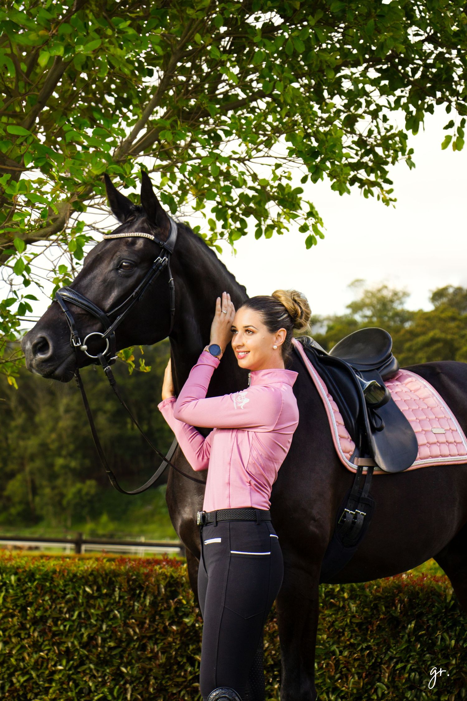 A person in pink interacts affectionately with a large black horse wearing a saddle outdoors.