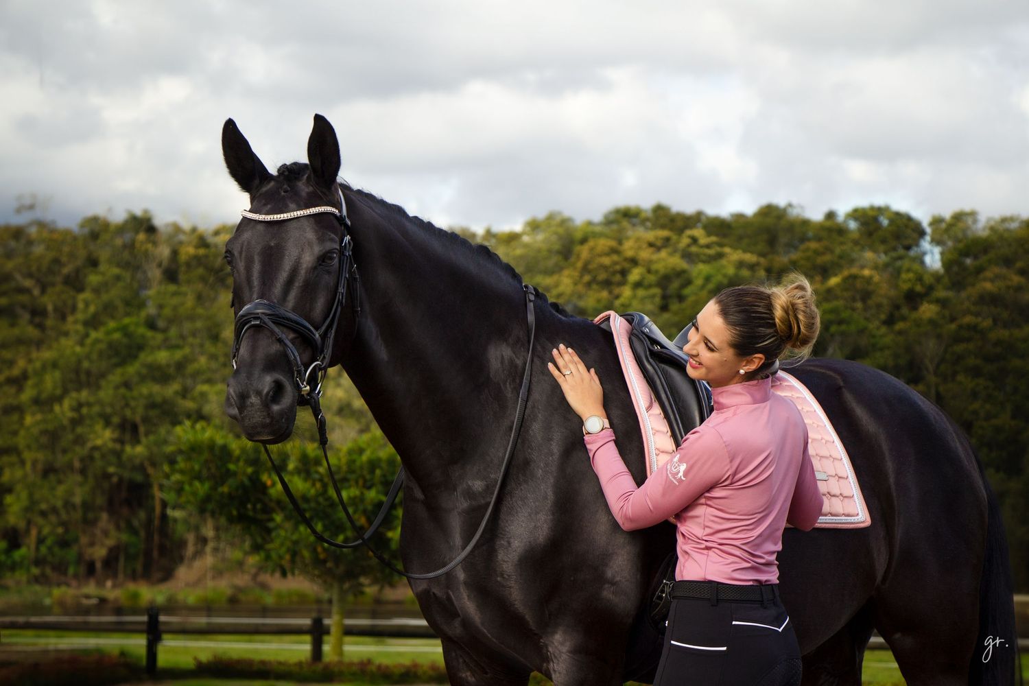 A rider in pink attire stands beside a black horse against a natural backdrop.