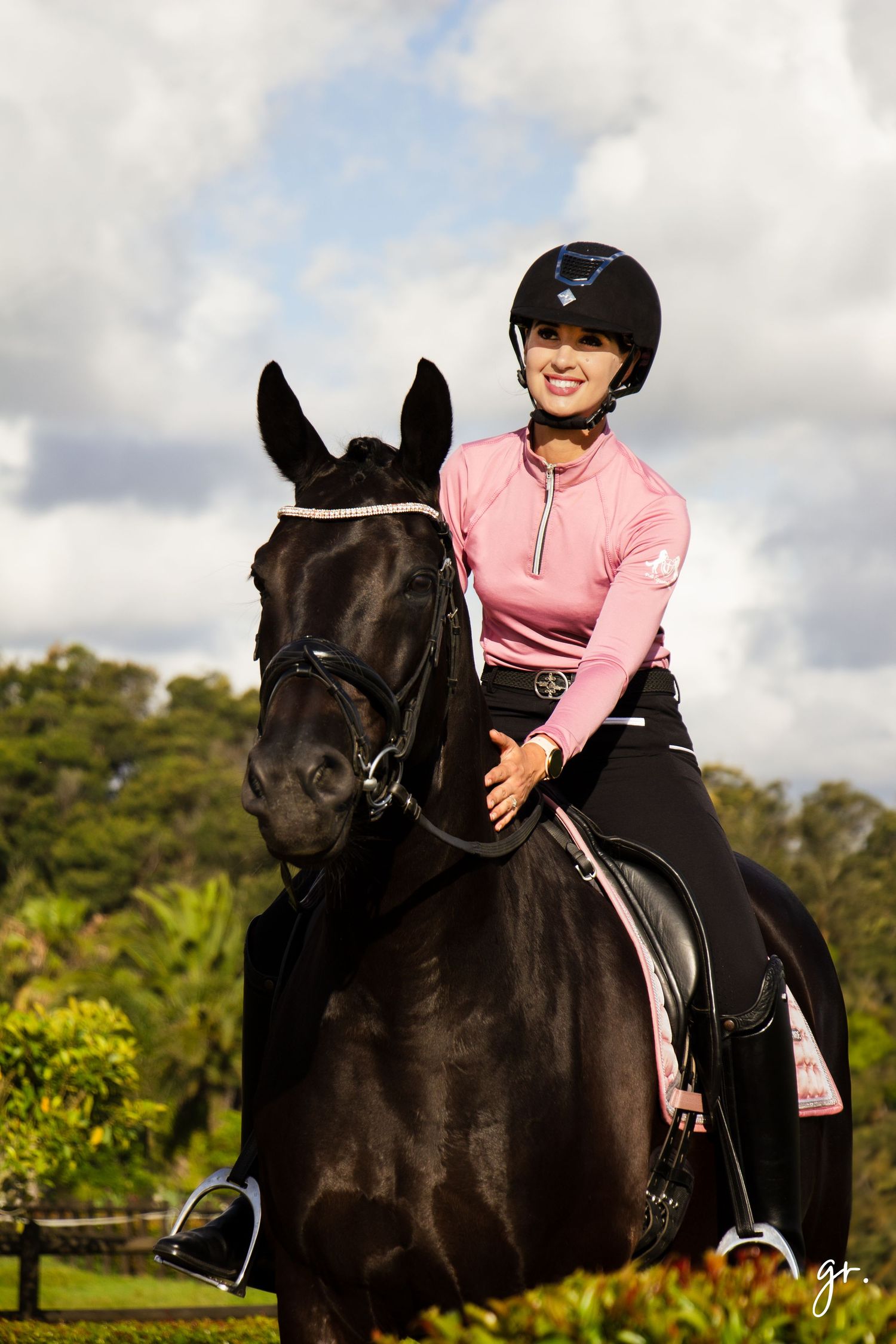 Black horse with rider in pink outfit against cloudy sky.