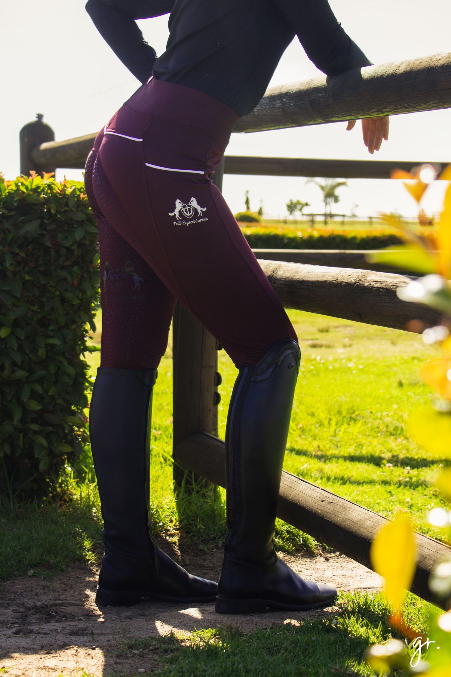 Purple riding breeches and boots shown near wooden fence in sunlit setting.