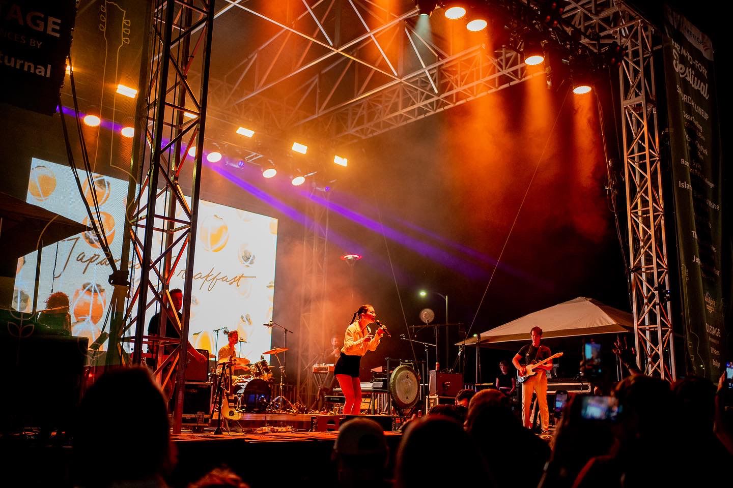 Concert stage illuminated with orange and purple lights during nighttime outdoor performance.