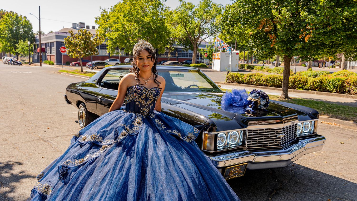 A flowing blue ball gown stands out against lush green trees and classic cars in an outdoor setting.