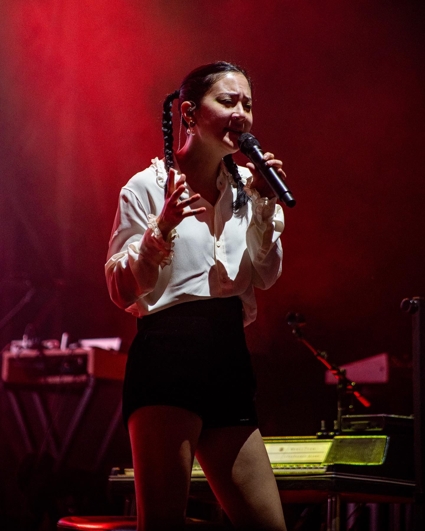 A performer in a white shirt and dark vest sings passionately on stage bathed in dramatic red lighting.