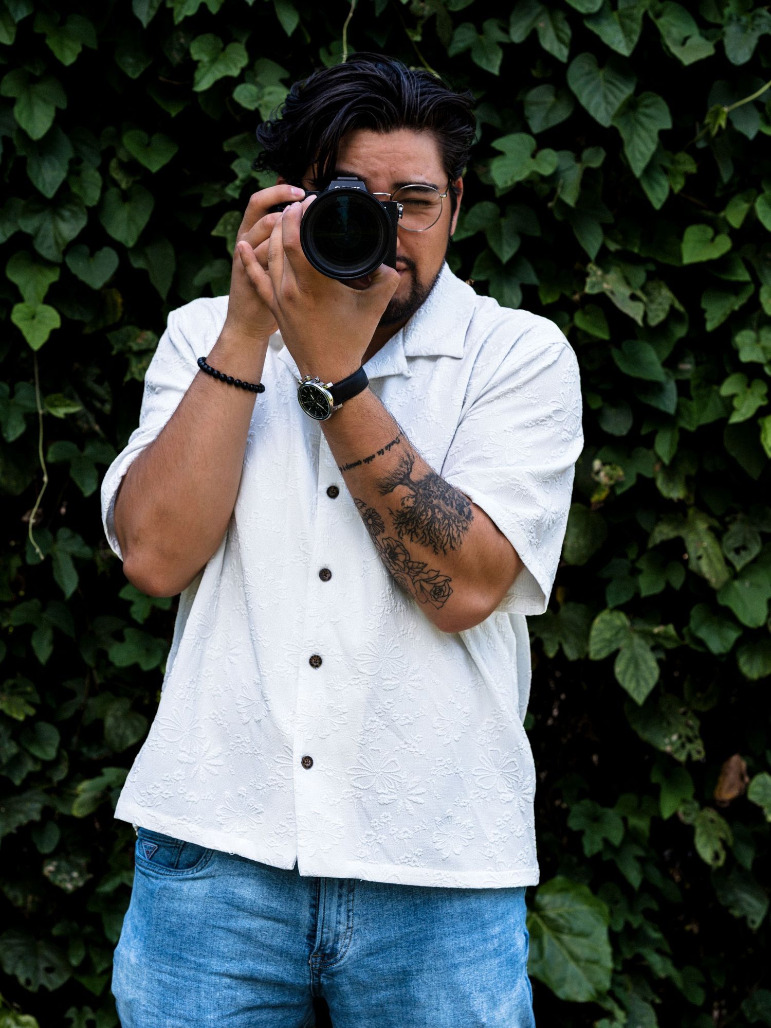 A photographer in a white shirt and jeans takes photos against a leafy green backdrop in a casual outdoor setting.