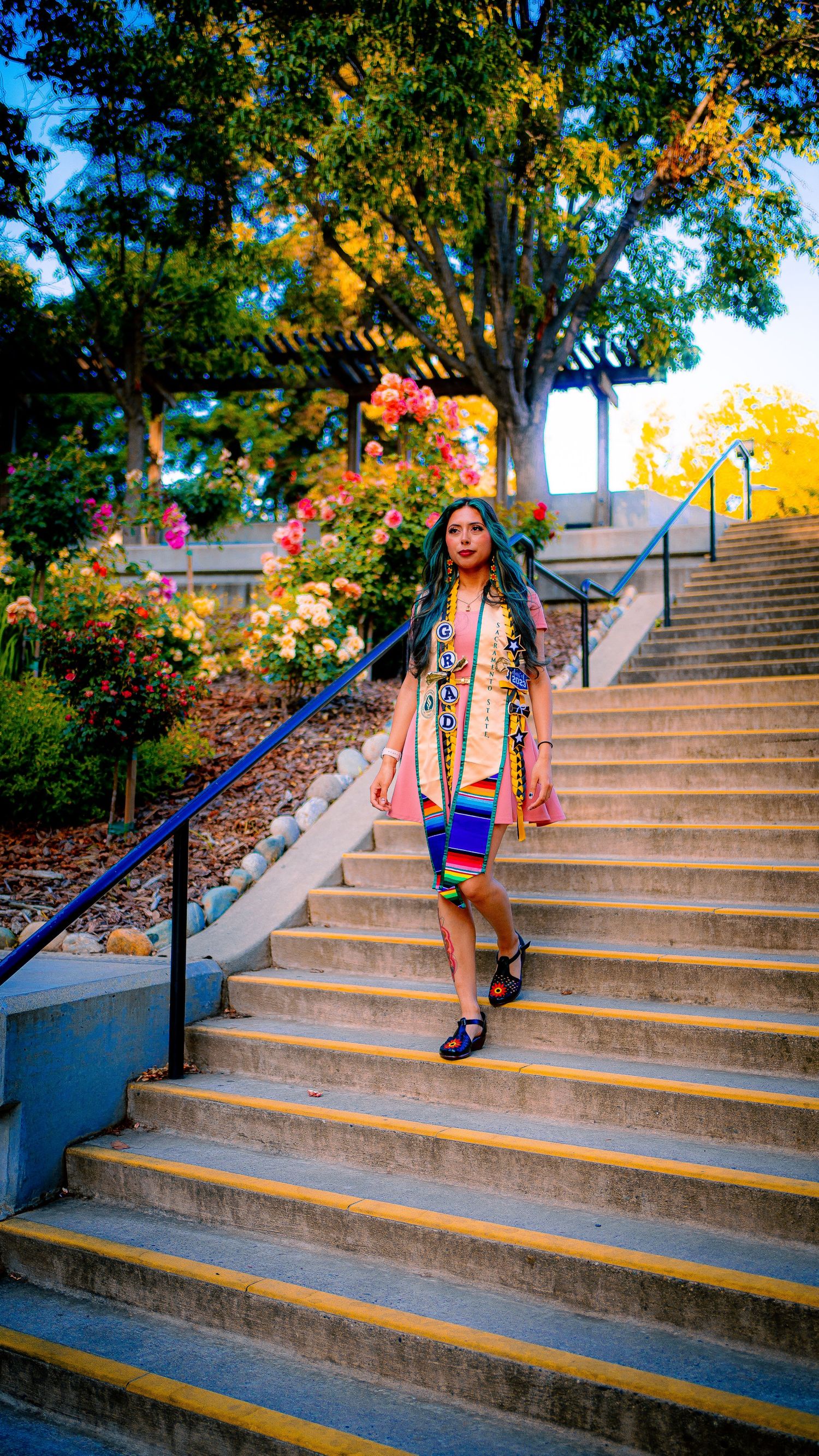 Graduate in regalia poses on outdoor campus stairs surrounded by autumn foliage.