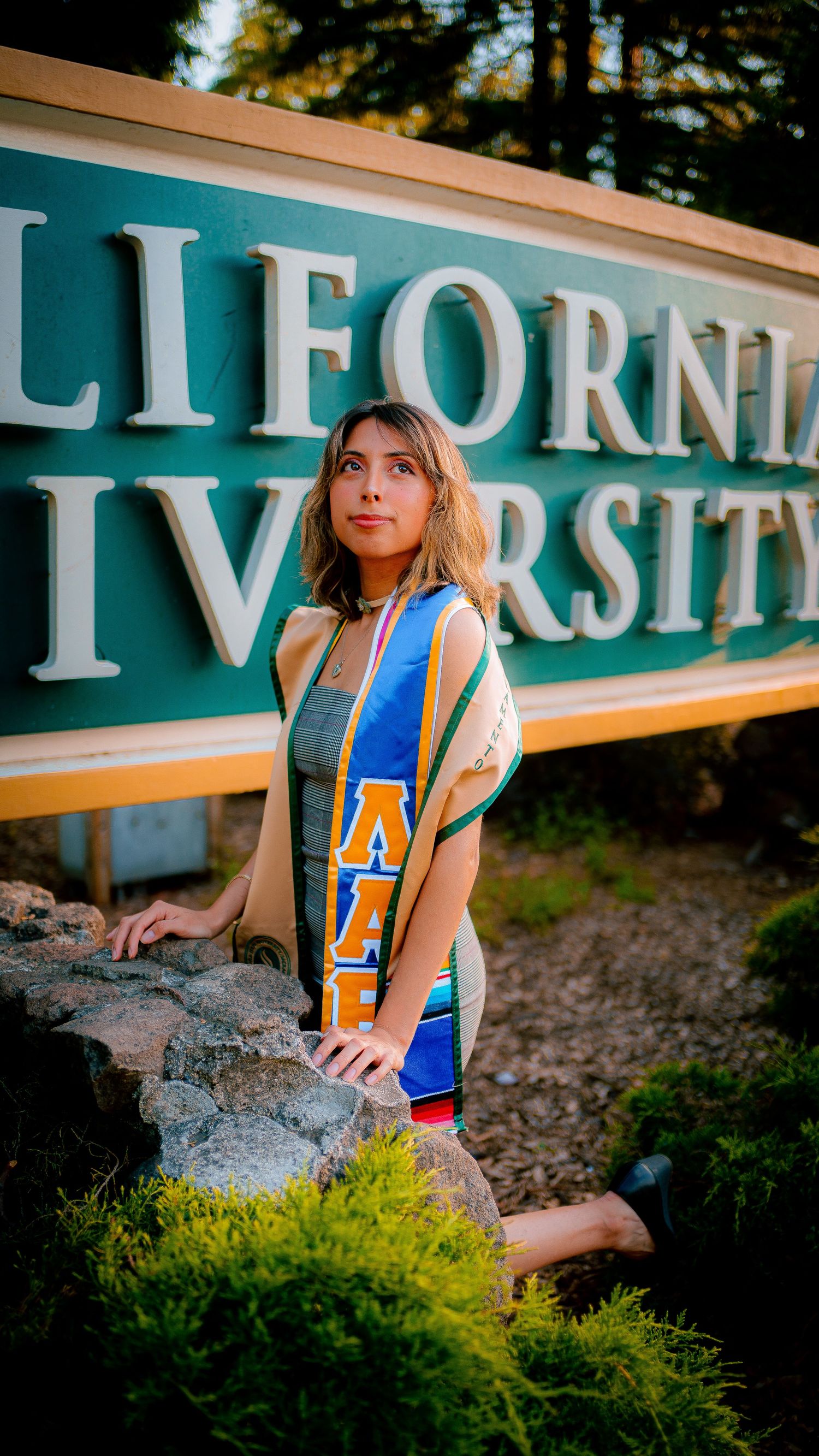 A person in a blue and yellow varsity jacket poses near a California sign during golden hour.