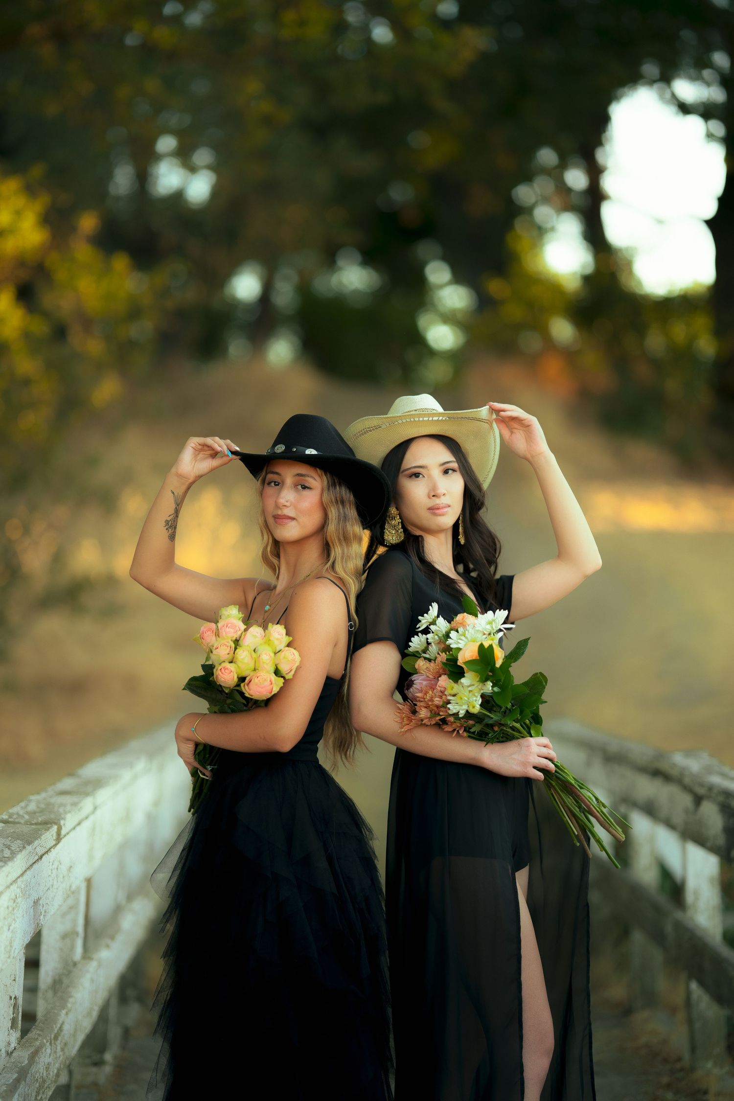 Two models in black dresses pose back-to-back on a bridge while holding colorful flower bouquets.
