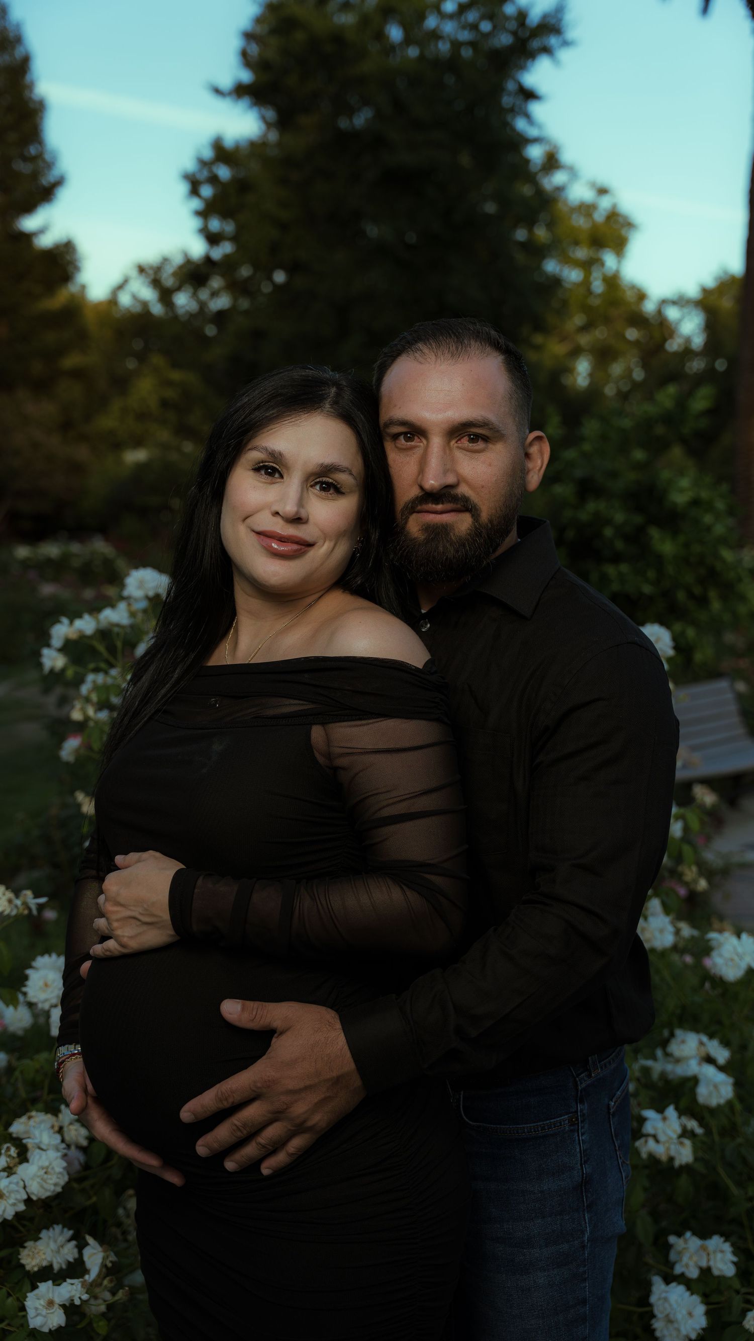 A couple in black attire embracing in an outdoor garden setting.