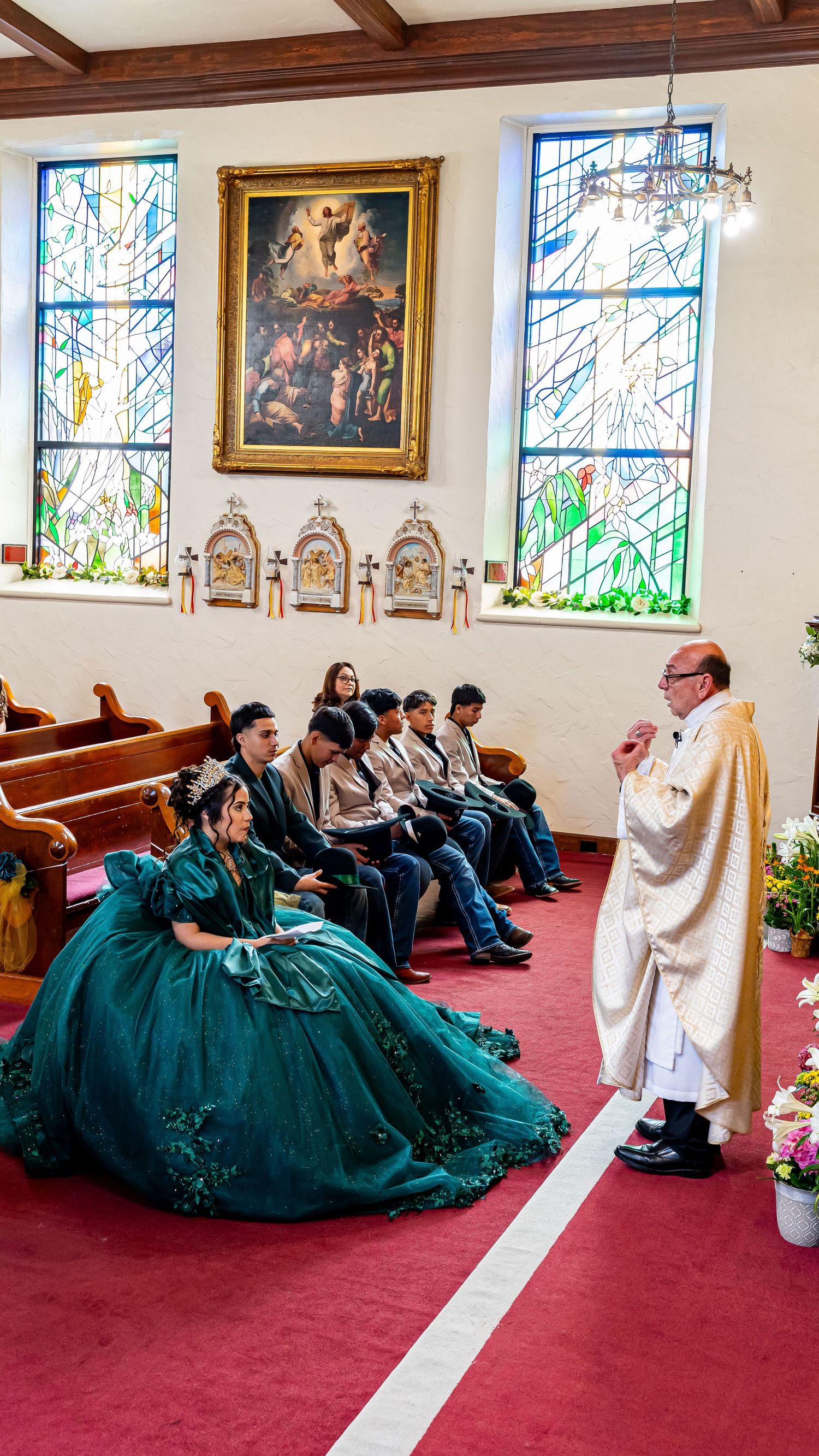 Traditional Catholic ceremony with clergy in white robes and decorative stained glass windows in background.