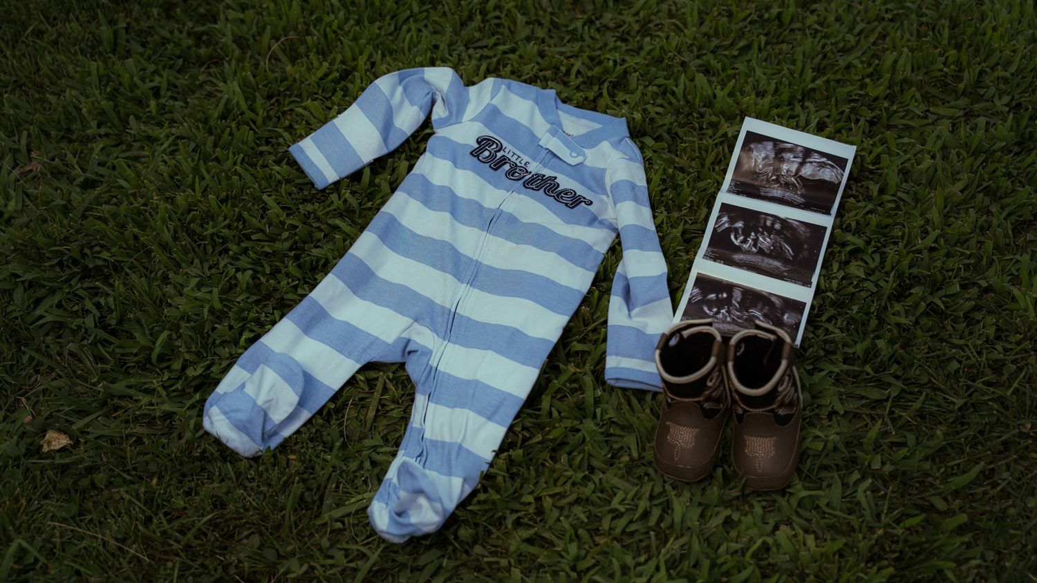 Baby items arranged on grass including striped onesie, boots and ultrasound photos.