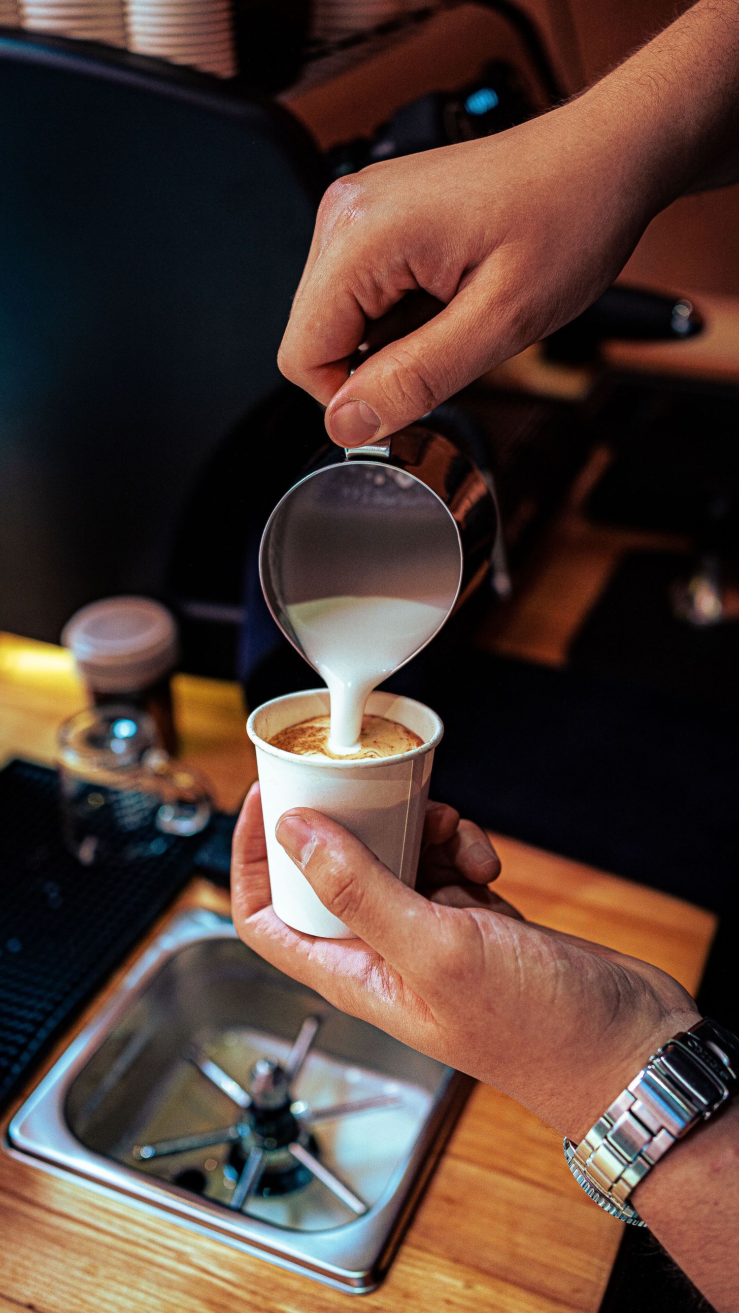 Hands pouring steamed milk into a coffee cup showing latte art preparation.