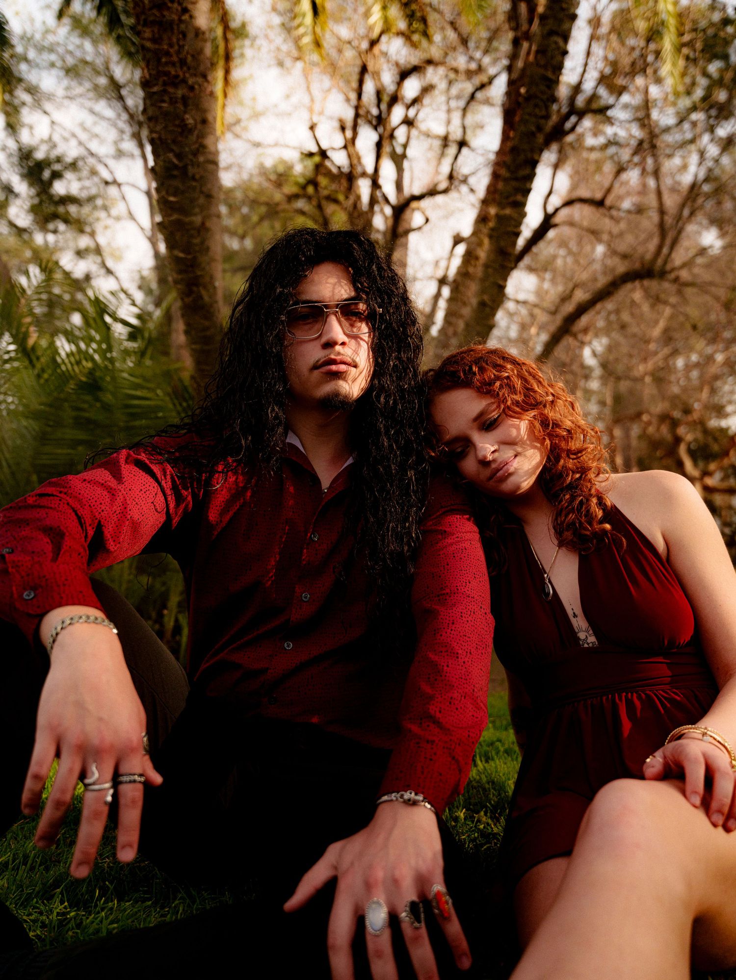 A couple in burgundy outfits sitting close together in a wooded outdoor setting during autumn.