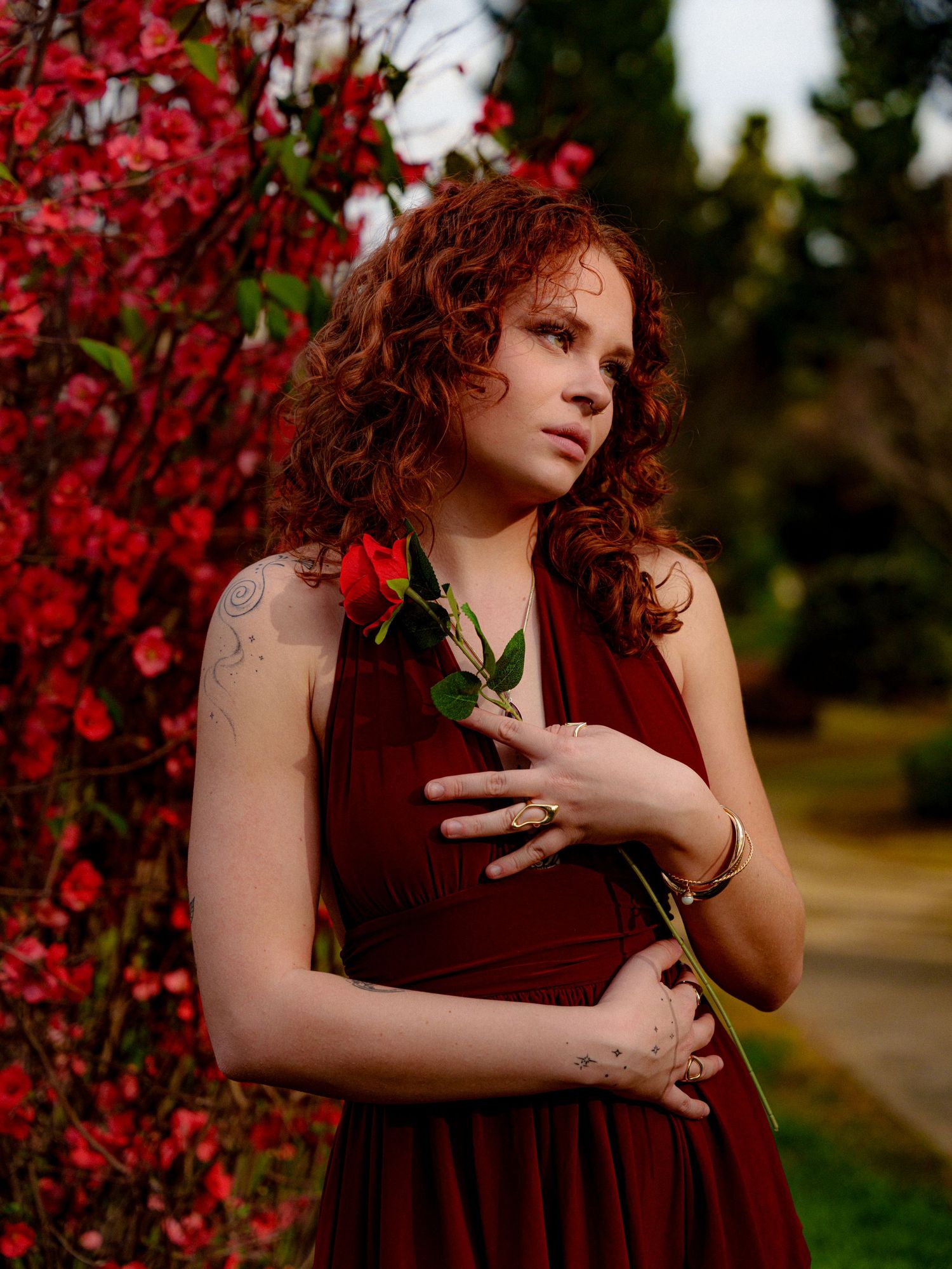 A figure in a burgundy dress poses gracefully against vibrant red spring blossoms in an outdoor setting.