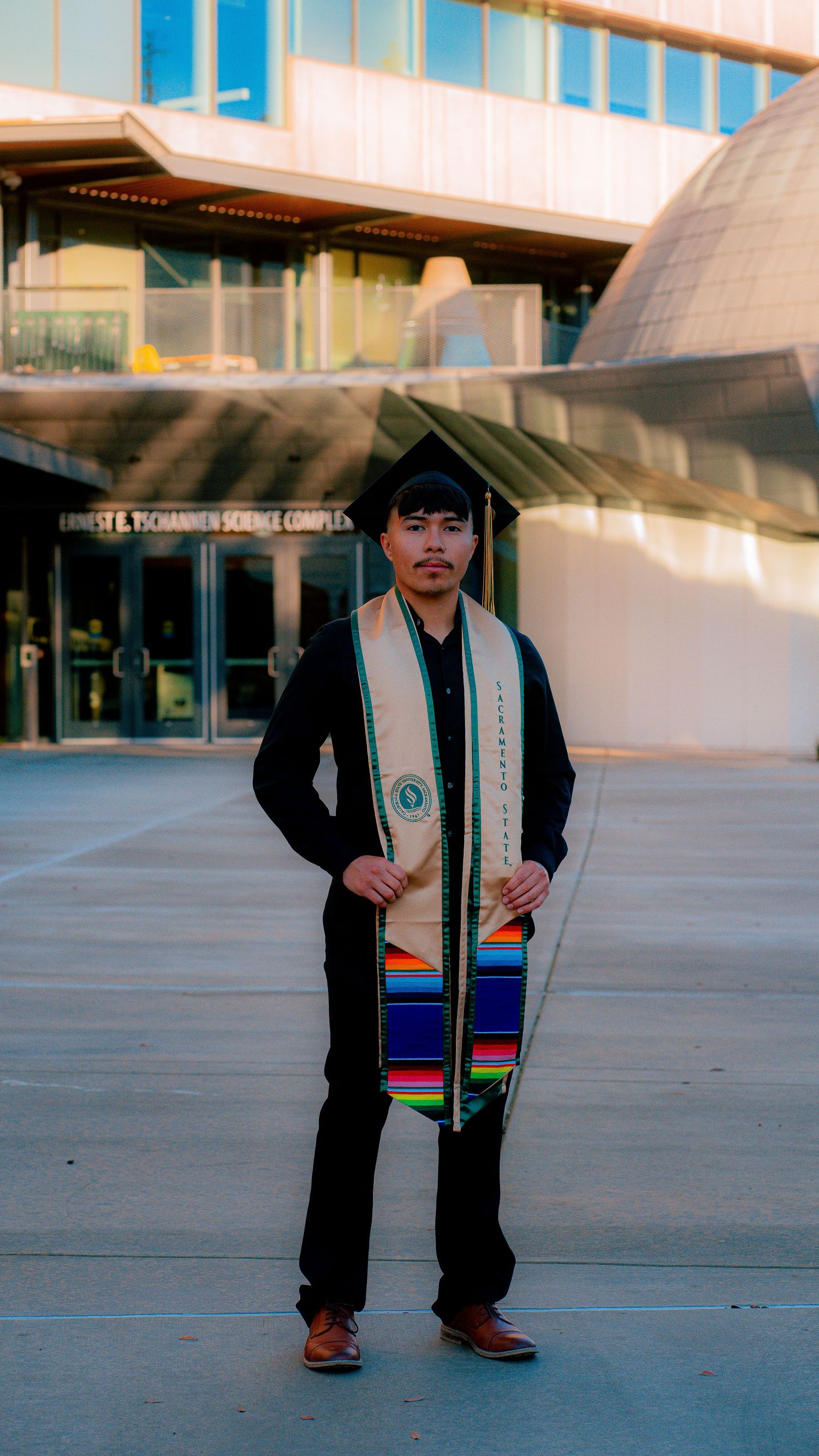 Graduate wearing stole and cap poses in front of modern campus architecture.