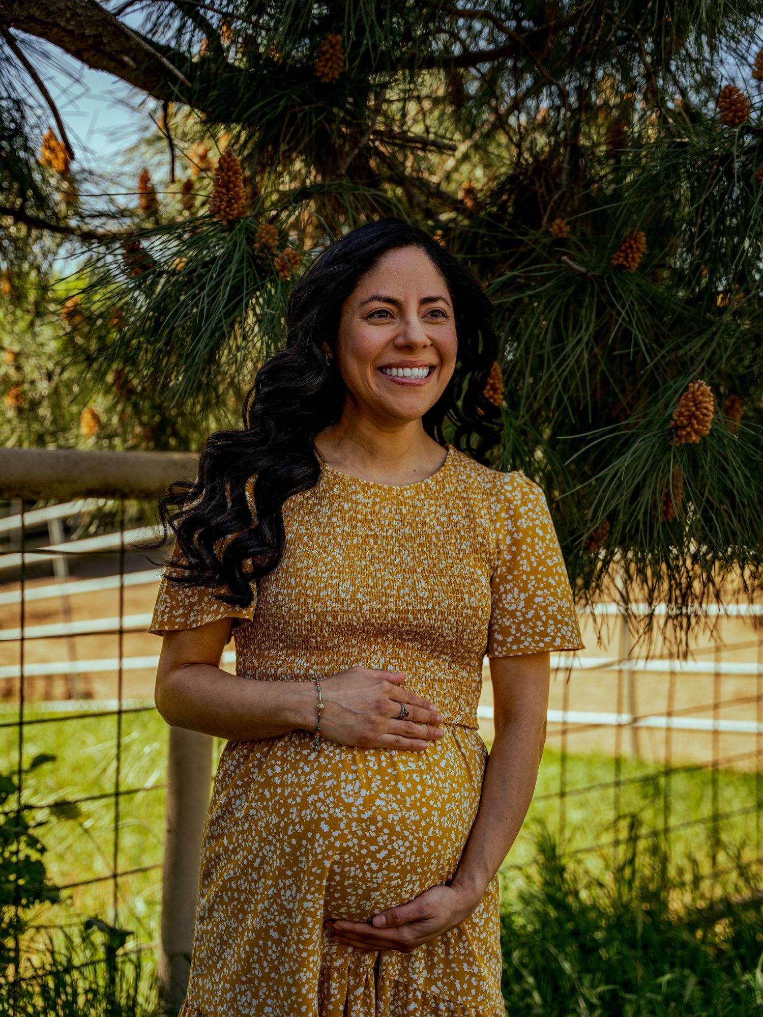 A person in a yellow floral dress poses outdoors against a natural backdrop with pine trees and autumn foliage.