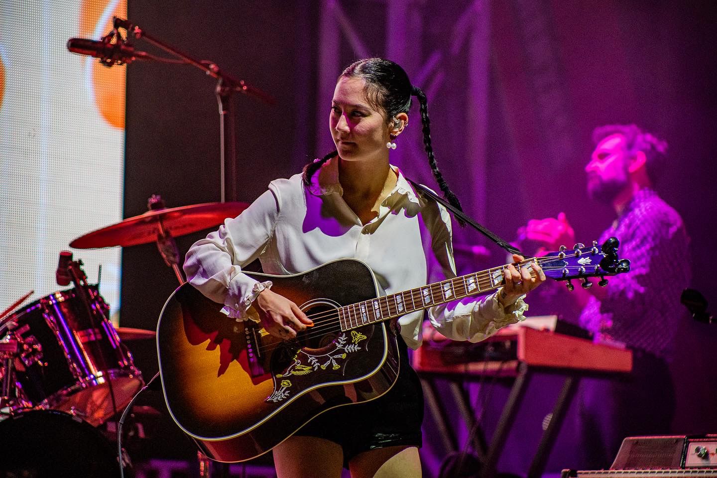 Musician performs on stage with acoustic guitar under purple lighting during live concert performance.
